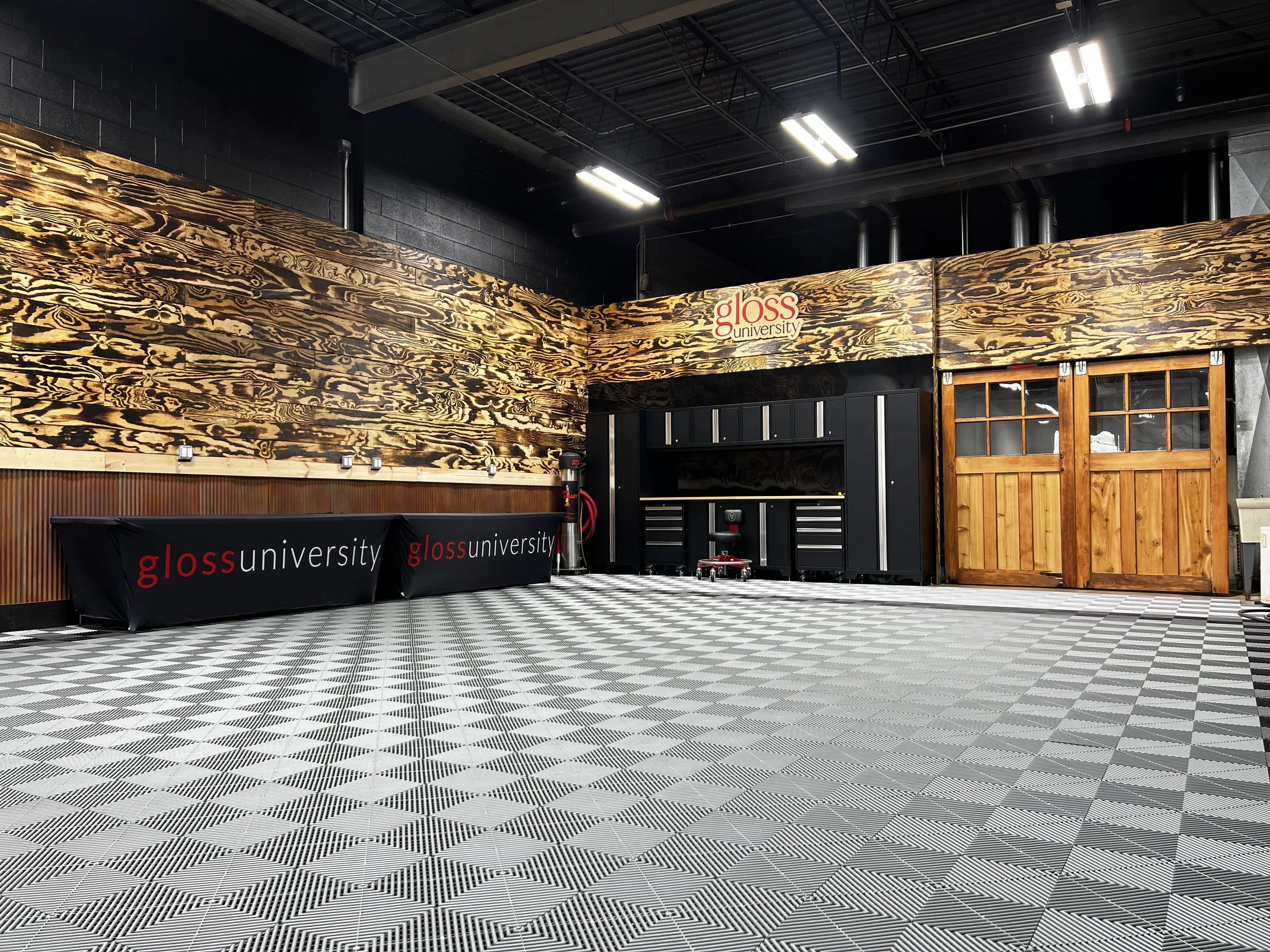 Interior of a room with black walls and wooden accents, featuring a black table with gloss university branding, a stack of black storage units, a fire extinguisher, a large wooden sliding door, and a checkered floor.