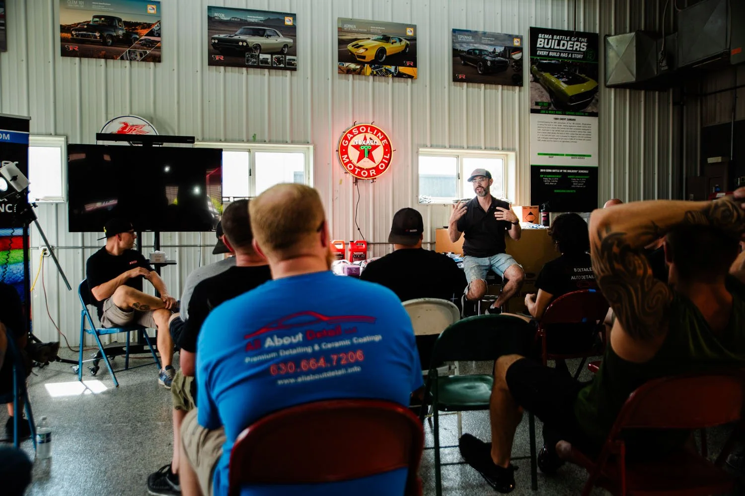 A group of people attending a presentation or workshop inside a garage or automotive building with car posters on the wall, a large TV, and a neon Gasoline sign. The presenter, a man with a beard, glasses, and a cap, is speaking to the audience.
