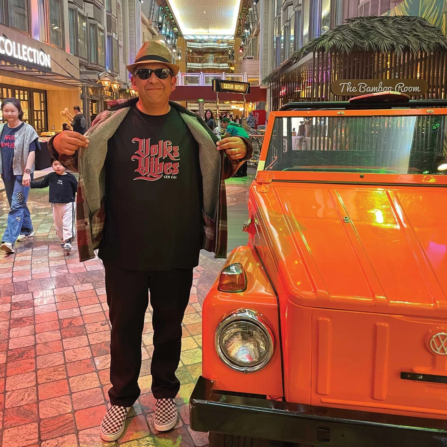 Man wearing sunglasses, a hat, and checkered shoes standing next to an orange vintage Volkswagen vehicle inside a shopping mall or entertainment complex.