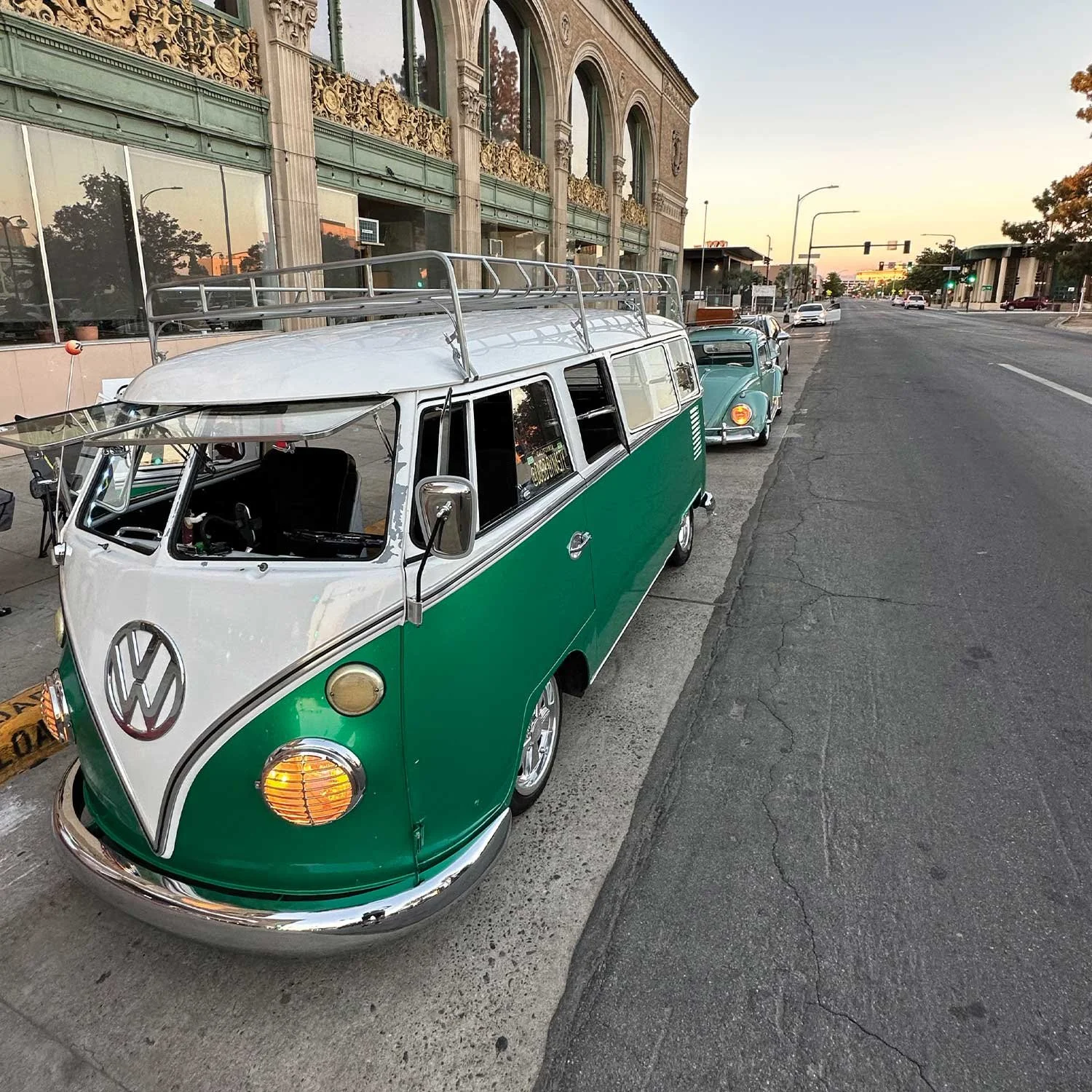 A row of vintage Volkswagen Type 2 (Microbus) vans parked along a city street sidewalk at sunset. The front van is green and white with a roof rack, and the others are light blue.