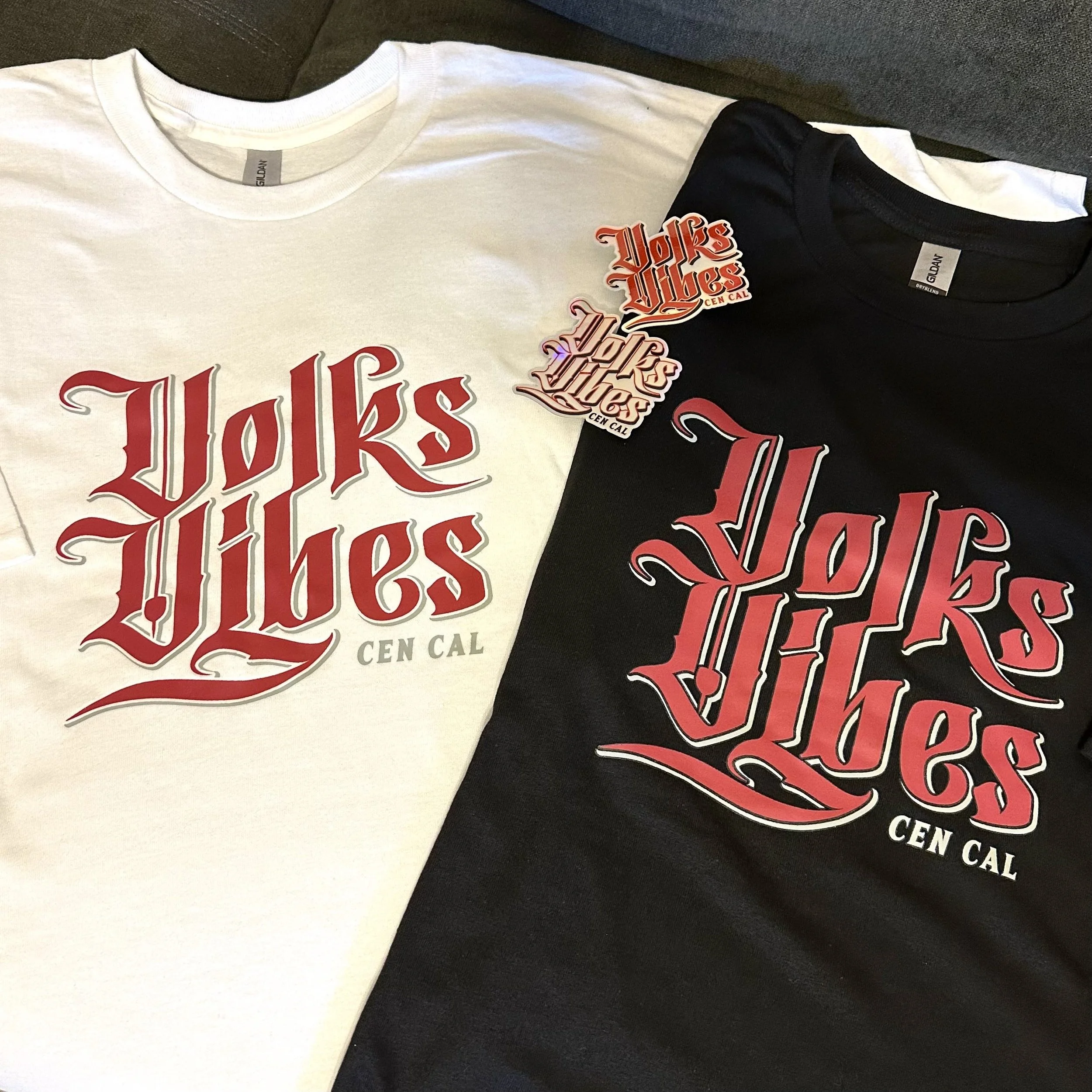 Two T-shirts, one white and one black, both with 'Voks Vibes CEN CAL' in stylized red and gray gothic font printed on the front. The T-shirts are laid flat, with two matching stickers placed on the white shirt.