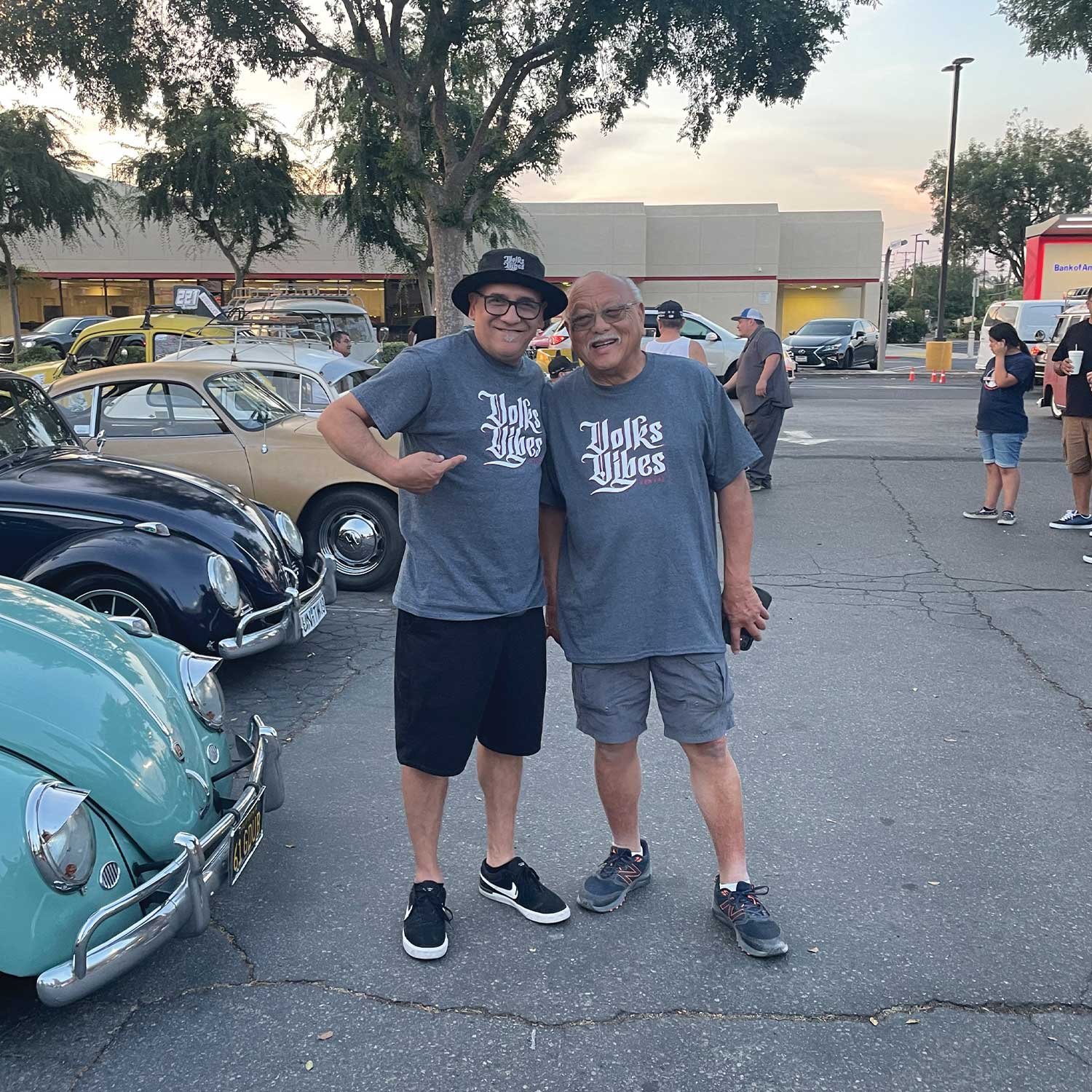 Two men wearing matching gray t-shirts with the words 'Volks Life' printed on them, standing in a parking lot among vintage cars, posing for the photo with smiles.