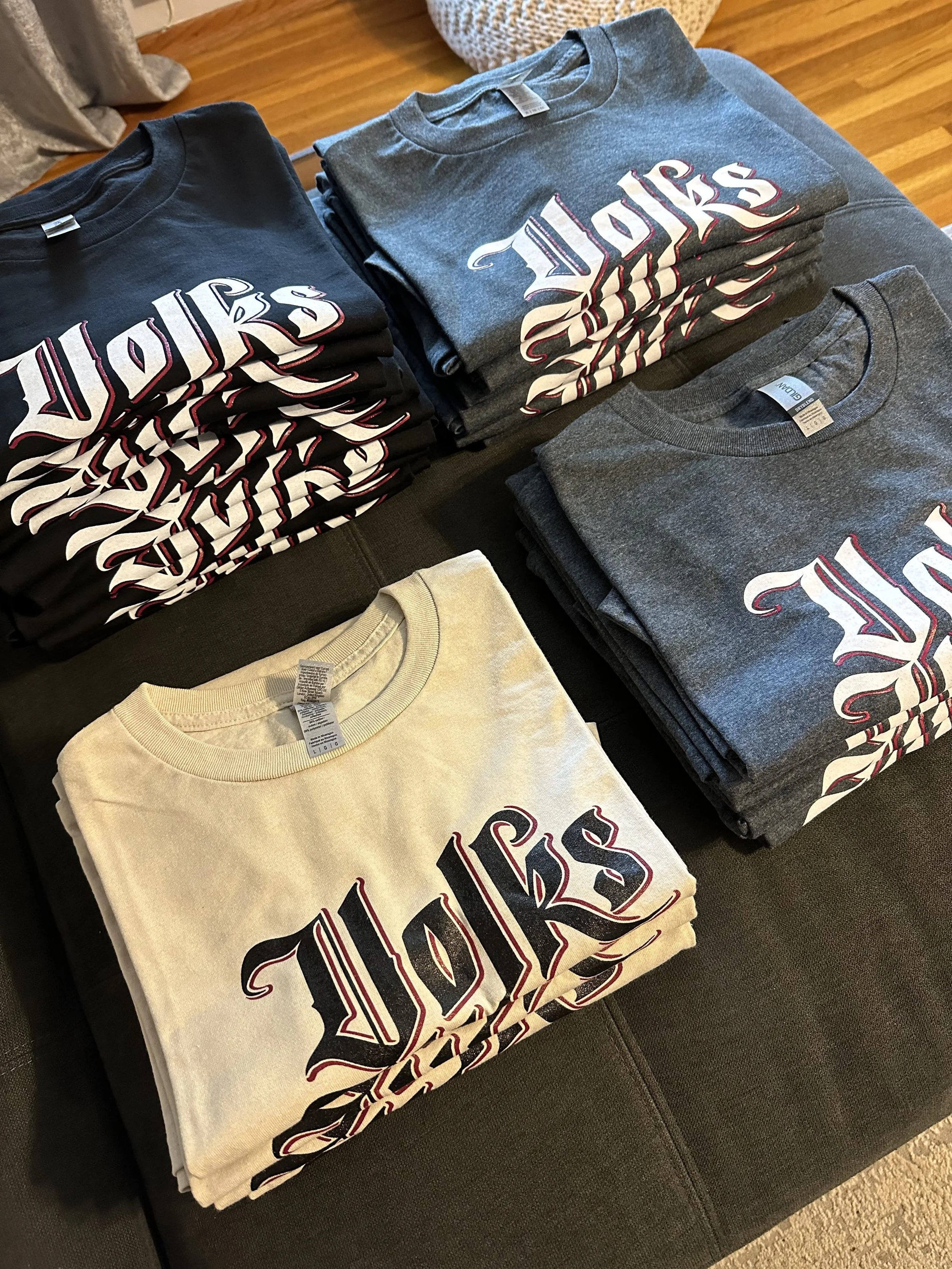 Stacks of T-shirts with the word 'VOLS' printed in a stylized font, arranged on a dark surface with a wooden floor in the background.