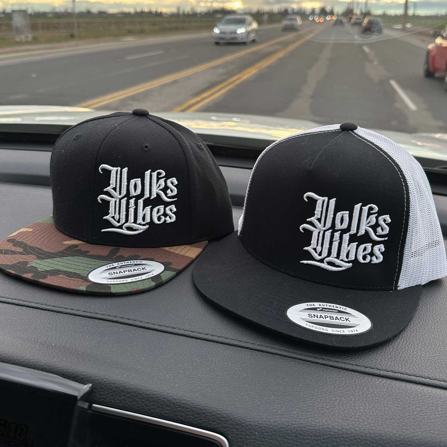 Two black and white trucker hats with the words 'Polks Vibes' embroidered on the front, placed on a car dashboard. One hat has a camouflage brim and the other has a plain black brim.