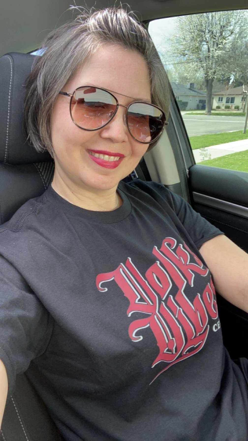 A woman with short gray hair, wearing sunglasses and a black T-shirt with red lettering, sitting inside a car and smiling.