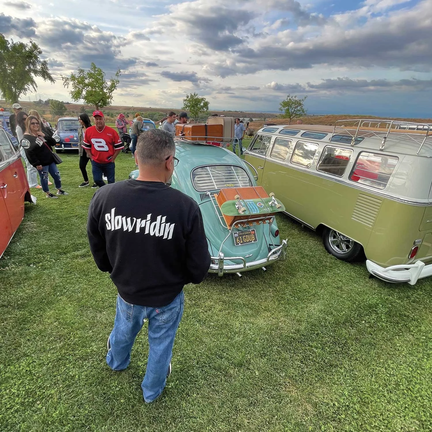 People gathered at a vintage car show on a grassy field under a partly cloudy sky, with several classic cars, including a blue and a green Volkswagen van, on display.