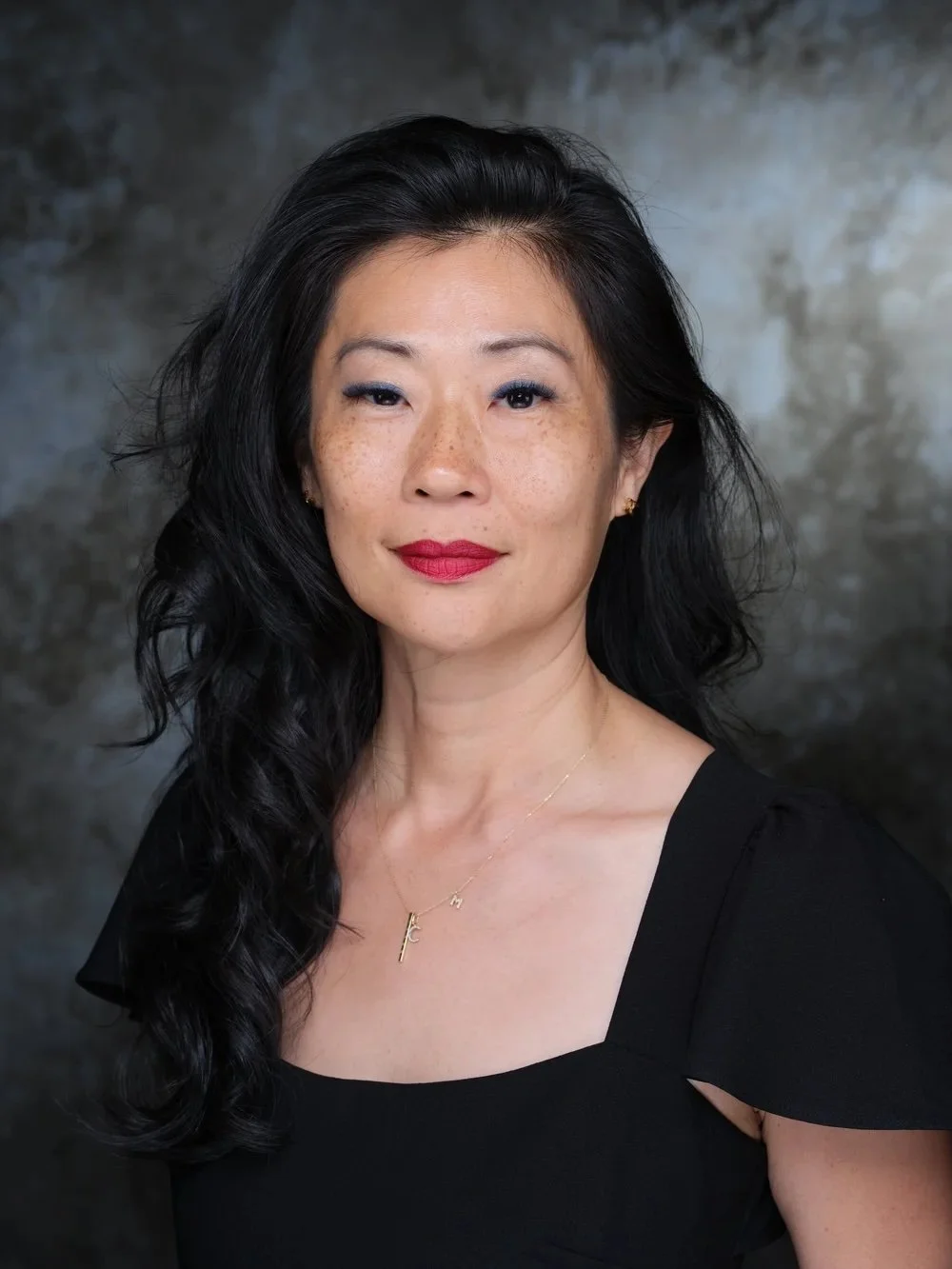 A middle-aged woman with wavy black hair and light skin, wearing red lipstick, a black top, and a gold necklace, posing against a dark grey mottled background.