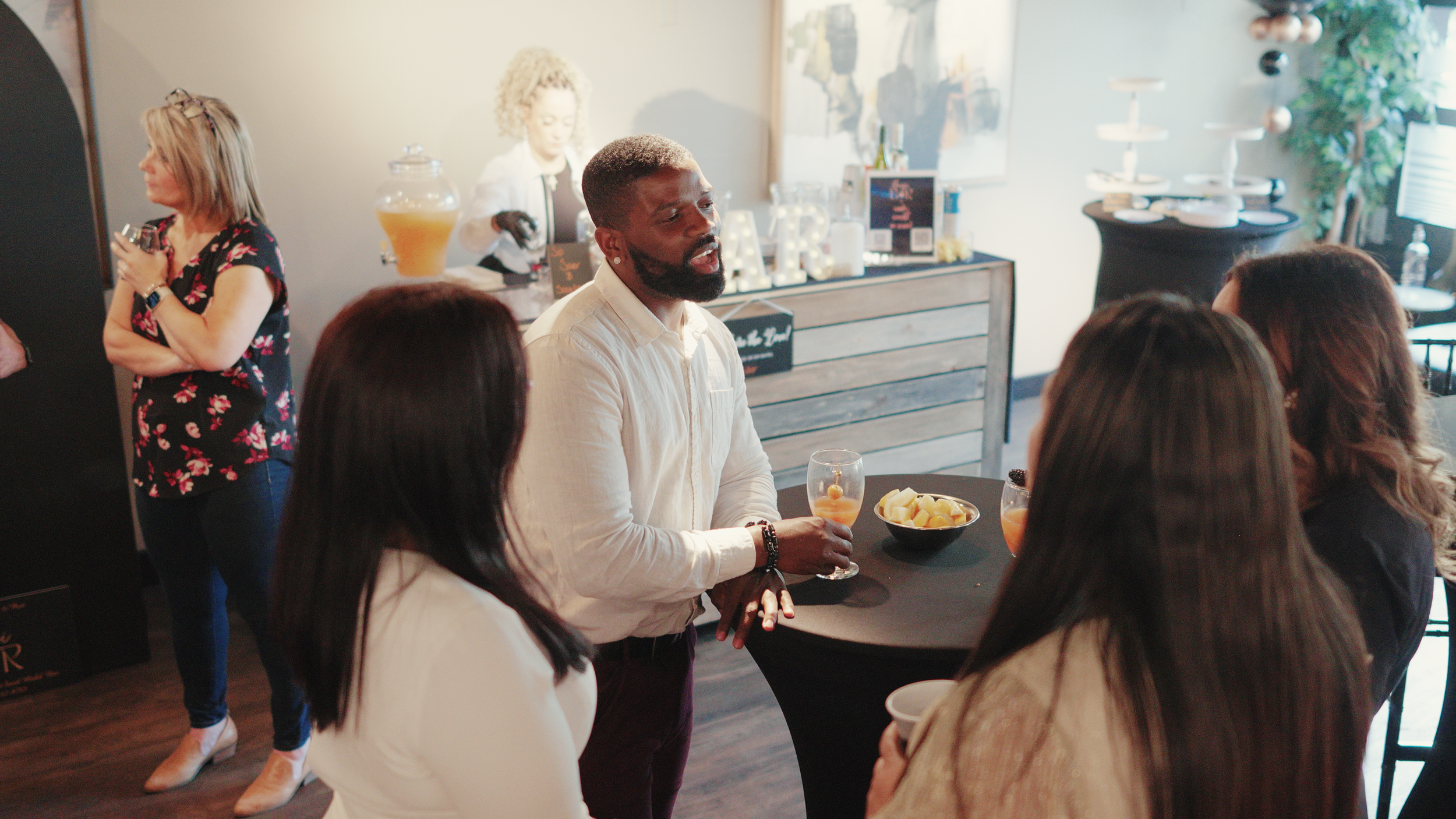 Social gathering with people mingling around a table, drinks in hand, an orange drink dispenser, and a fruit bowl on a table.