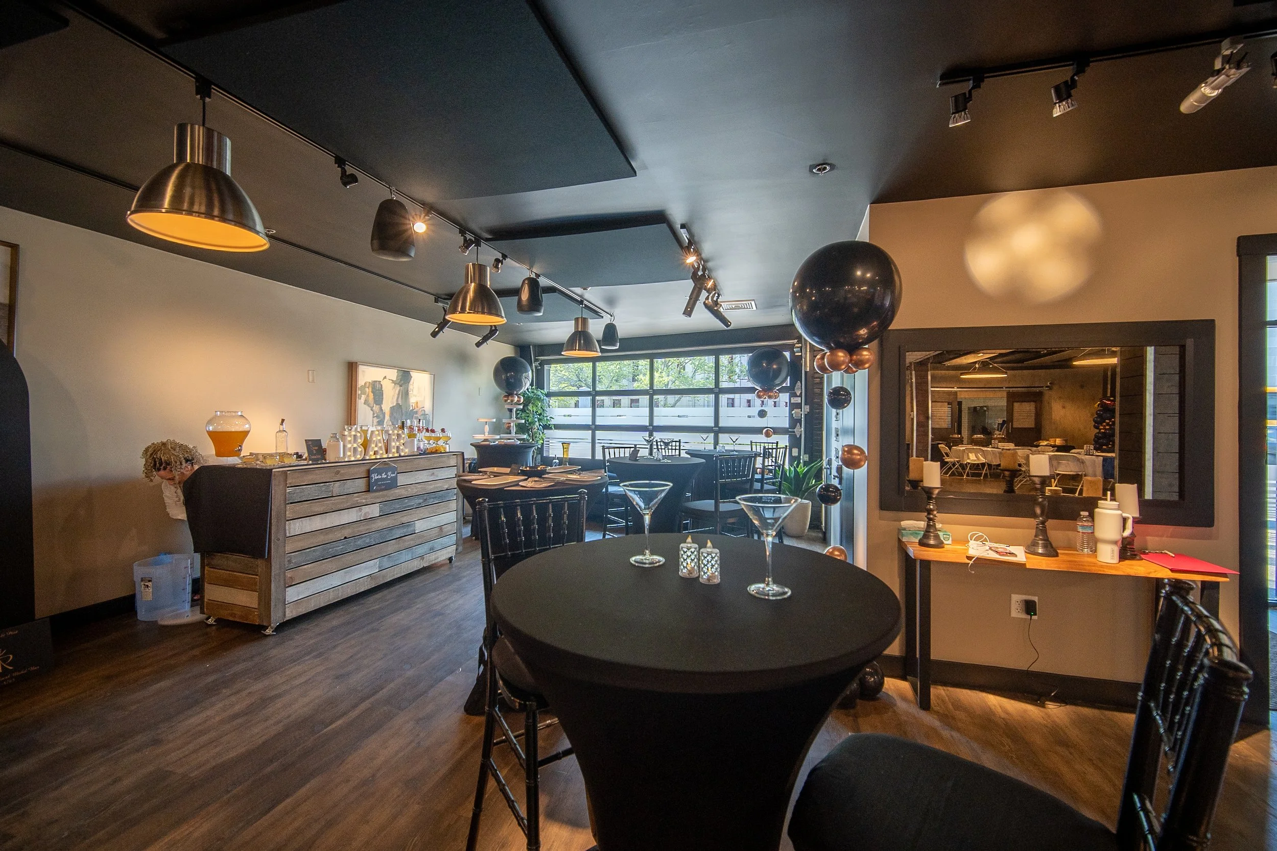 A decorated event space with black tablecloths, chairs, and balloons. A wooden bar is set up with drinks and decorations. Modern lighting fixtures hang from the ceiling, and large windows allow natural light. A large mirror is mounted on the wall, re