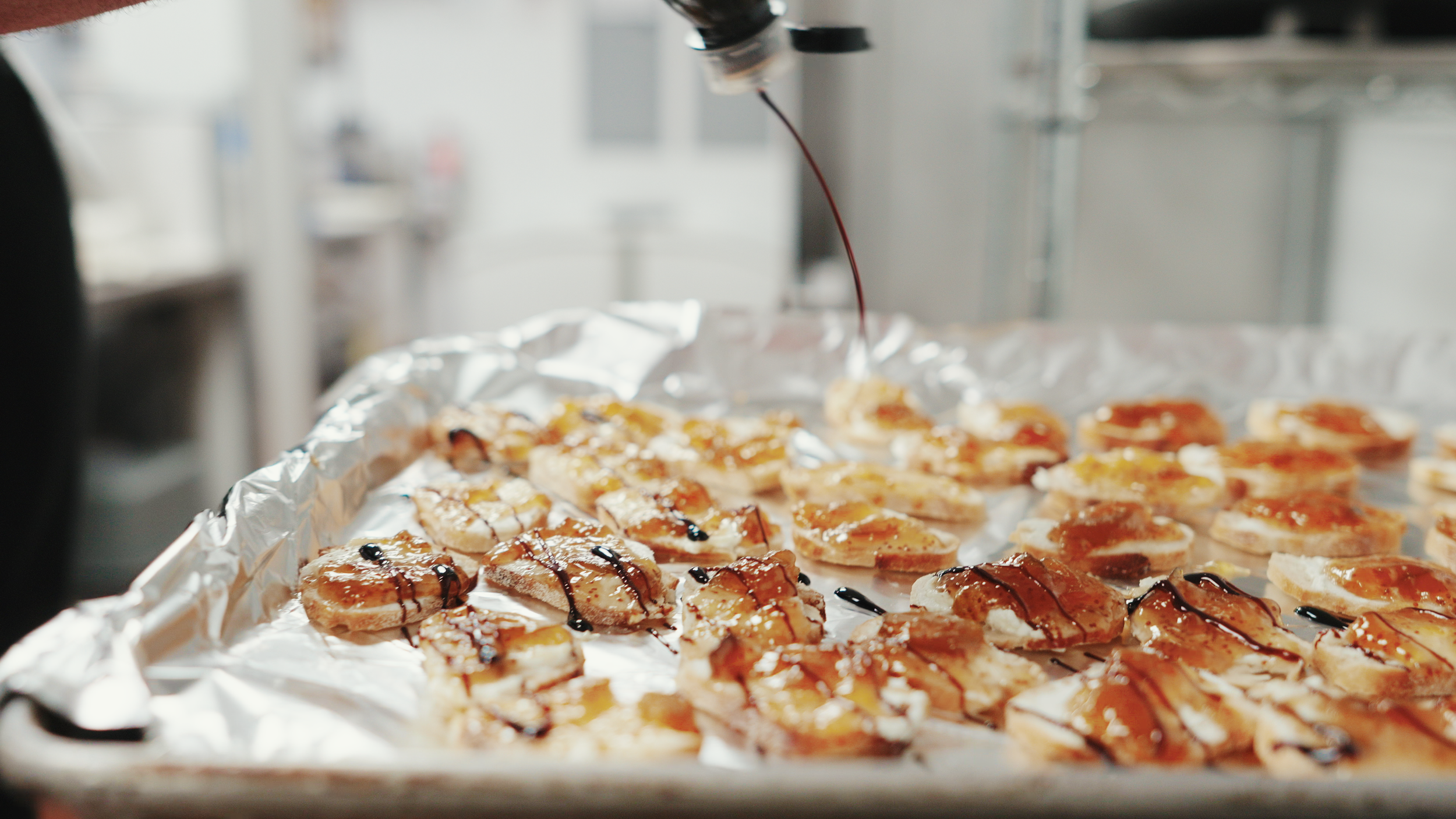 Sliced bread with cheese and jam, drizzled with balsamic glaze on foil-covered tray.