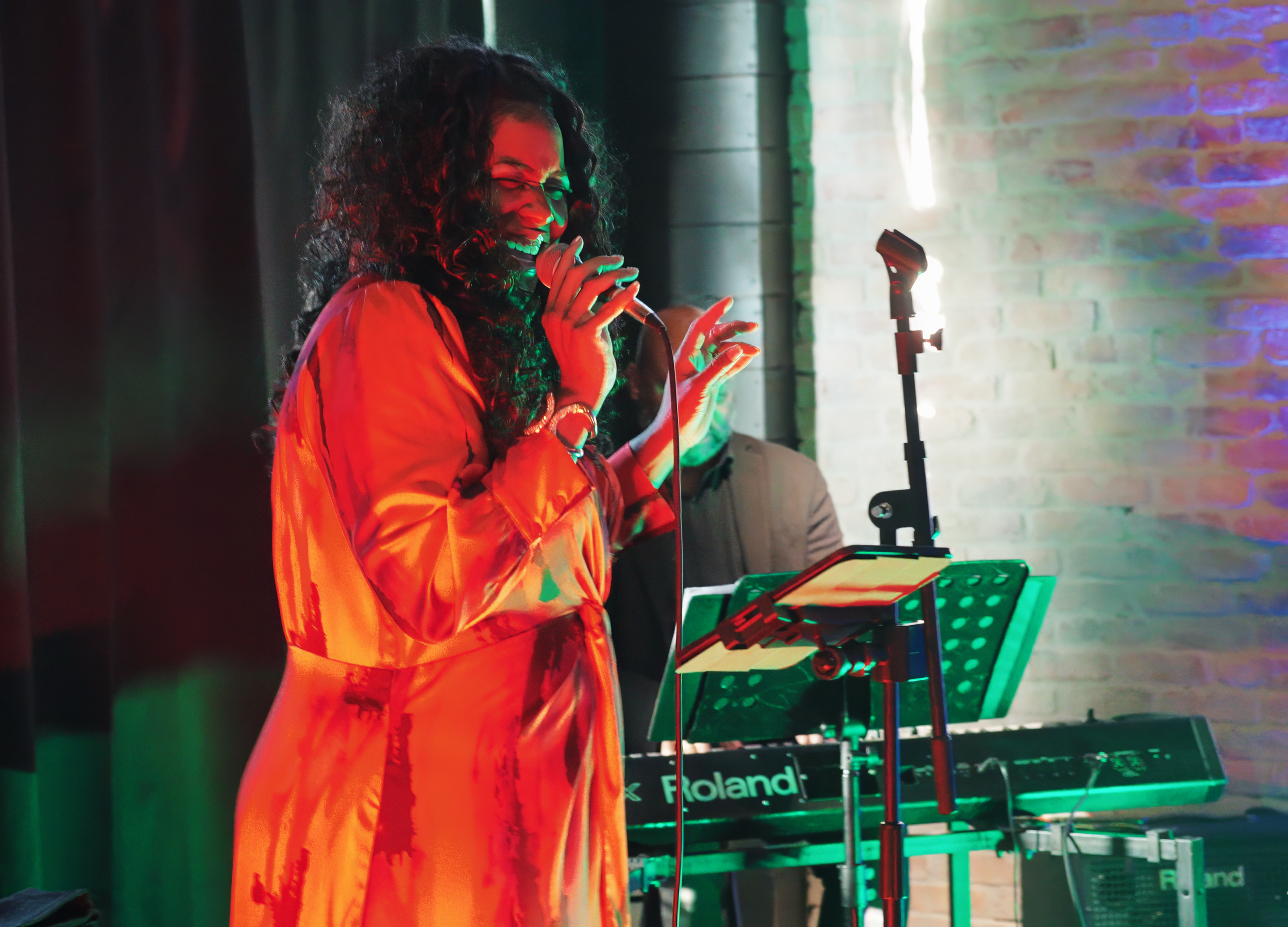 Woman singing into a microphone on stage with keyboard and music stand, colorful stage lighting, brick wall background.
