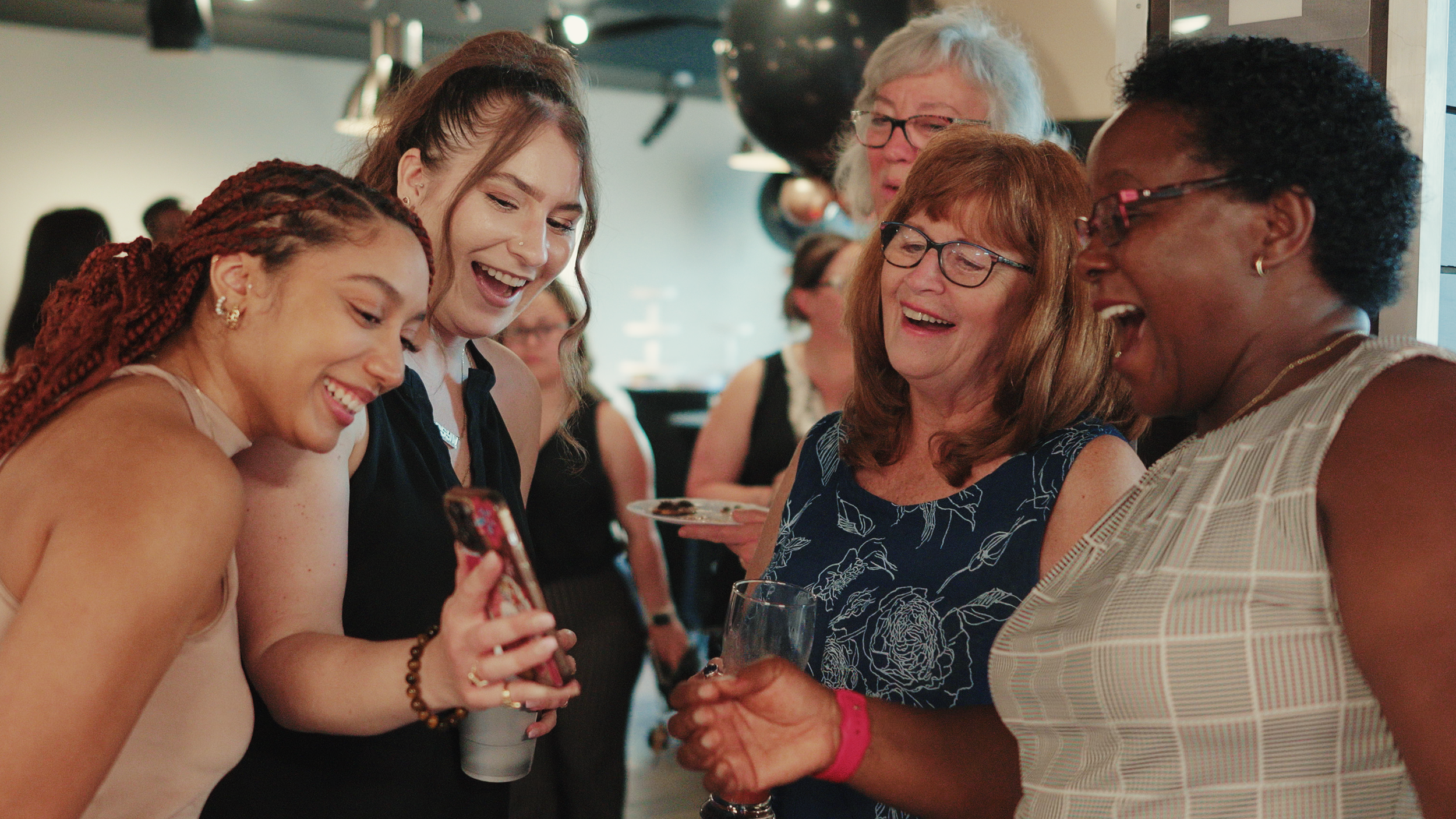 A group of diverse women smiling and looking at a smartphone together at a social gathering.