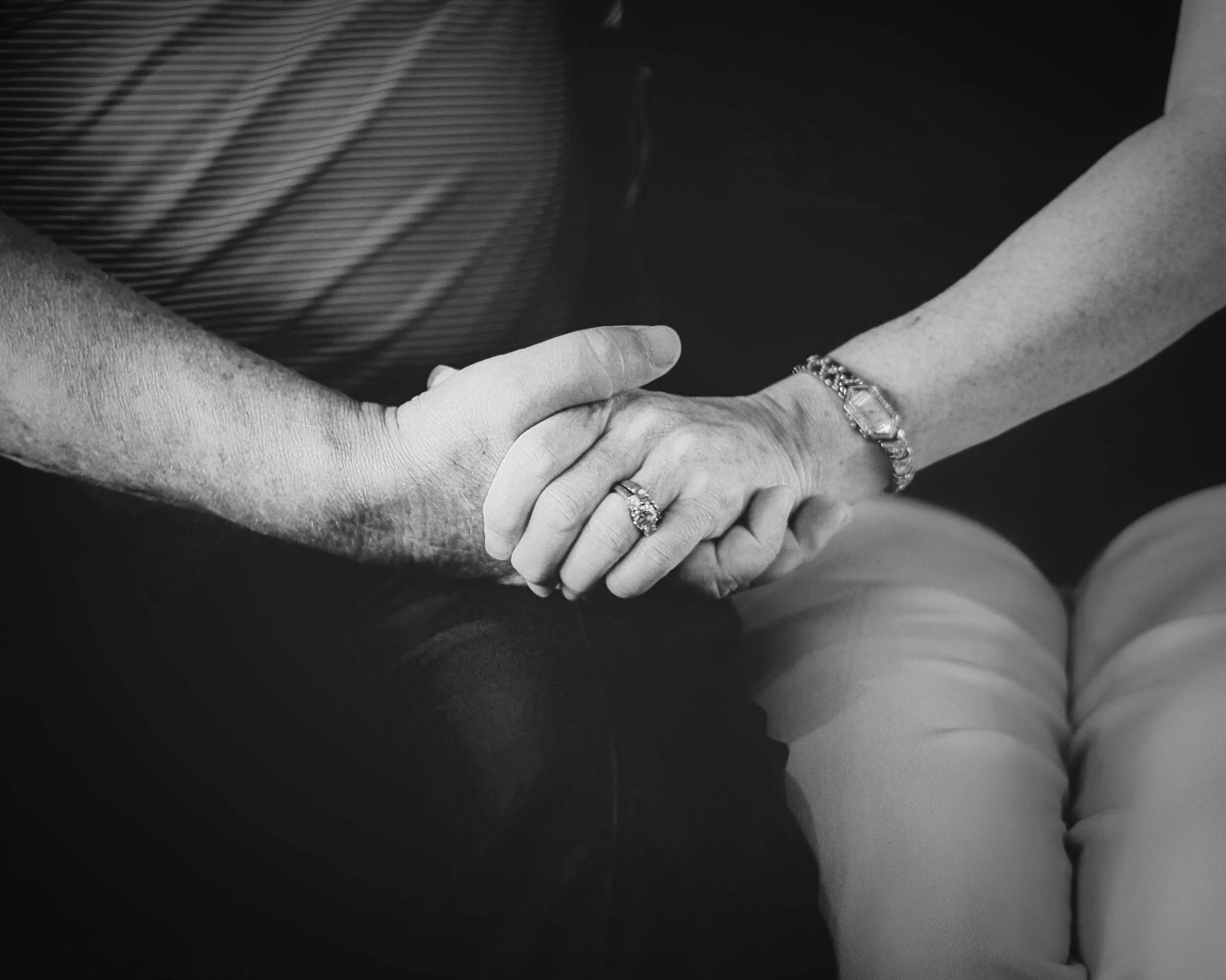 Close-up of an elderly couple holding hands, woman's hand adorned with a ring and bracelet, in black and white.