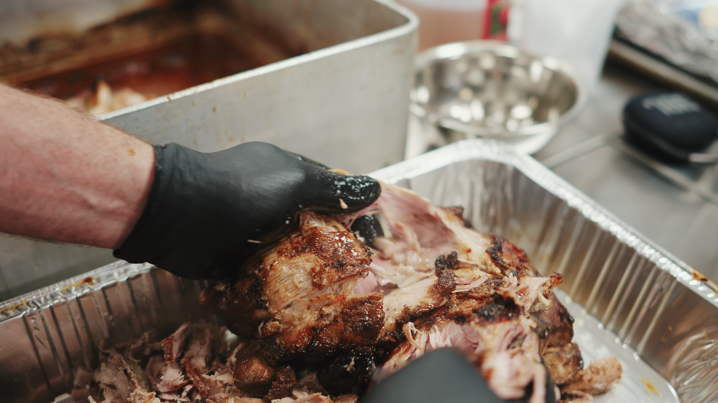 Person wearing black gloves pulling apart roasted meat in a metal tray.