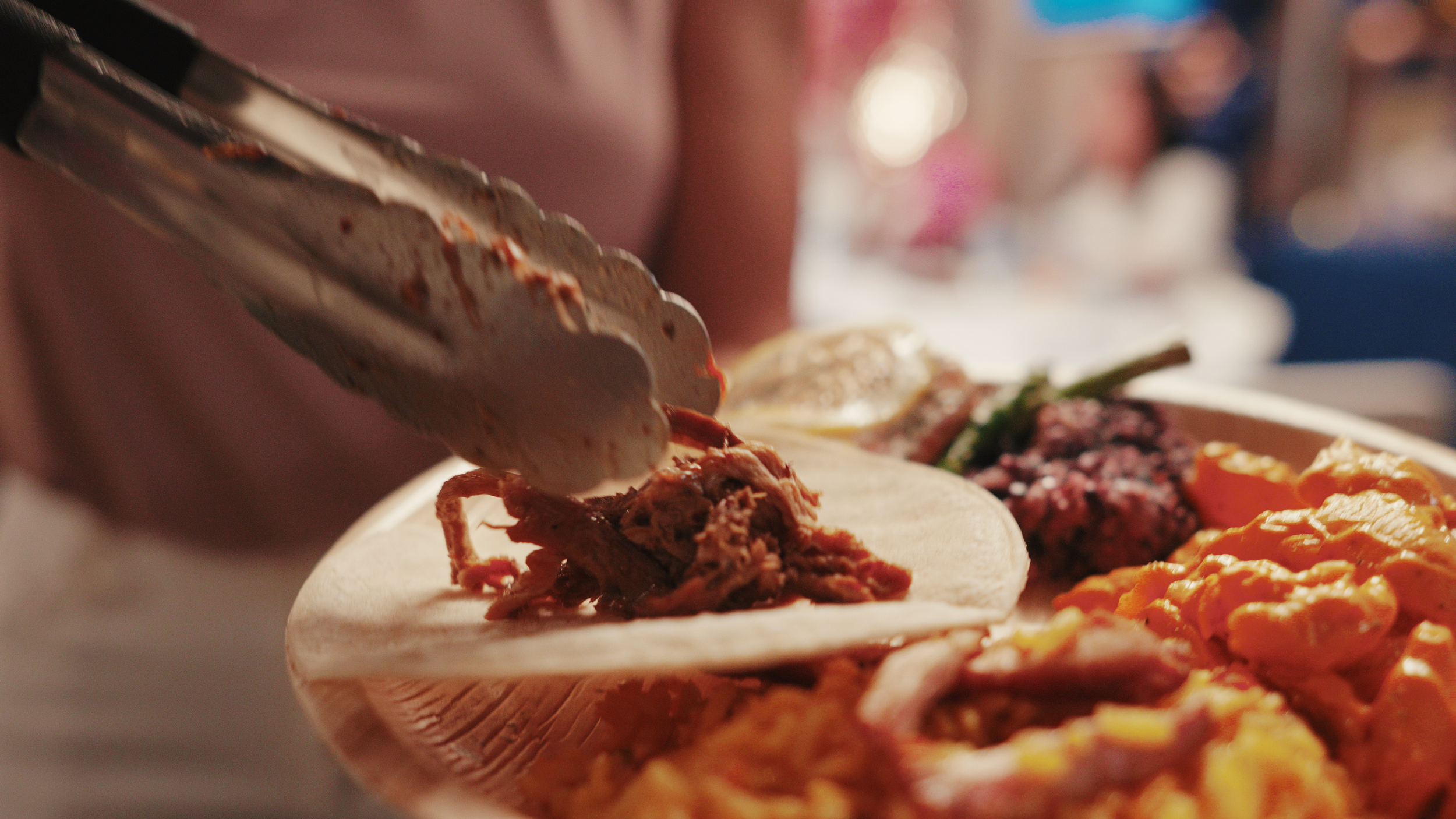 Tongs holding pulled meat over tortilla with plate of food including rice and vegetables