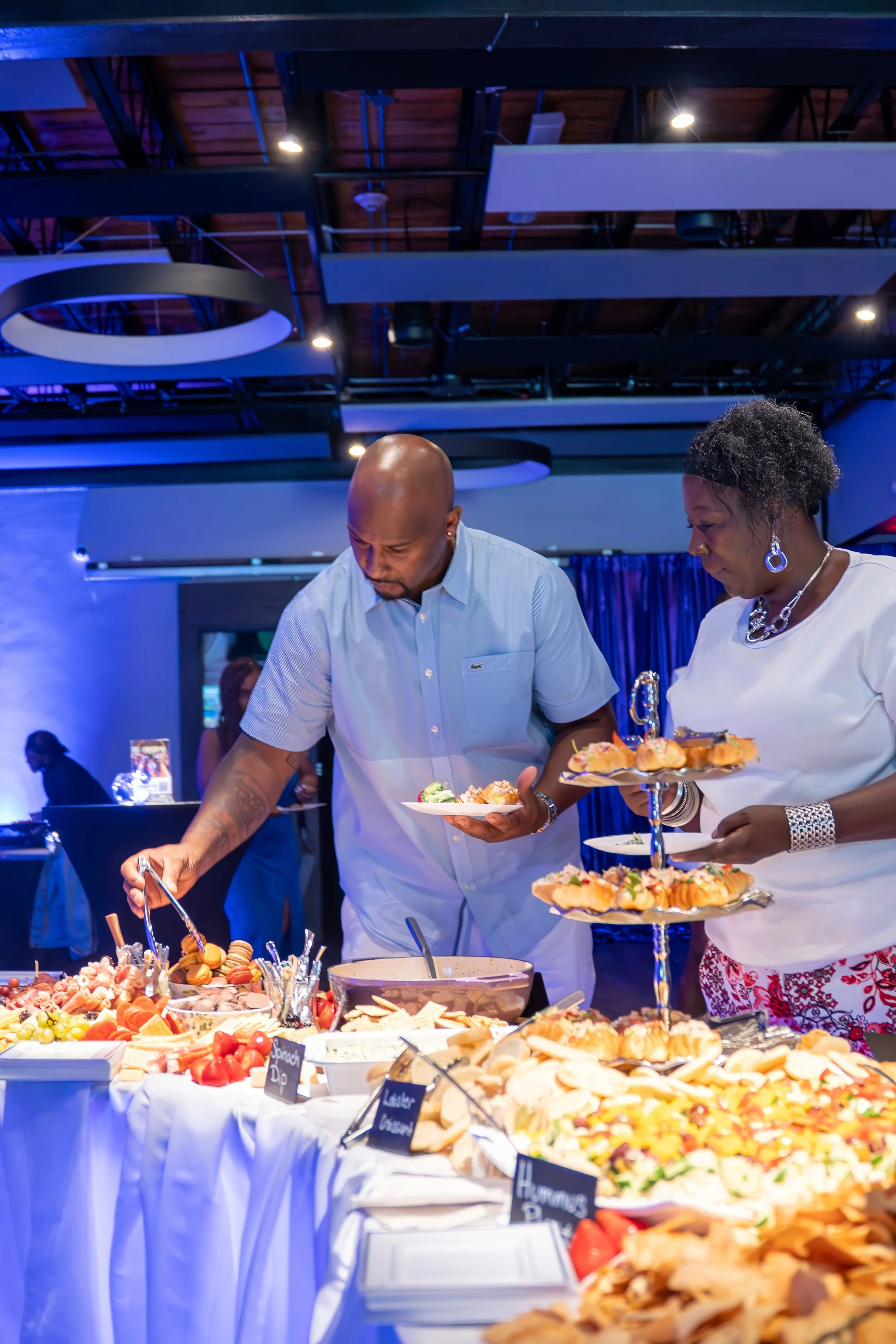 Two people serving themselves food from a buffet table at a party or event, with various appetizers and snacks displayed.