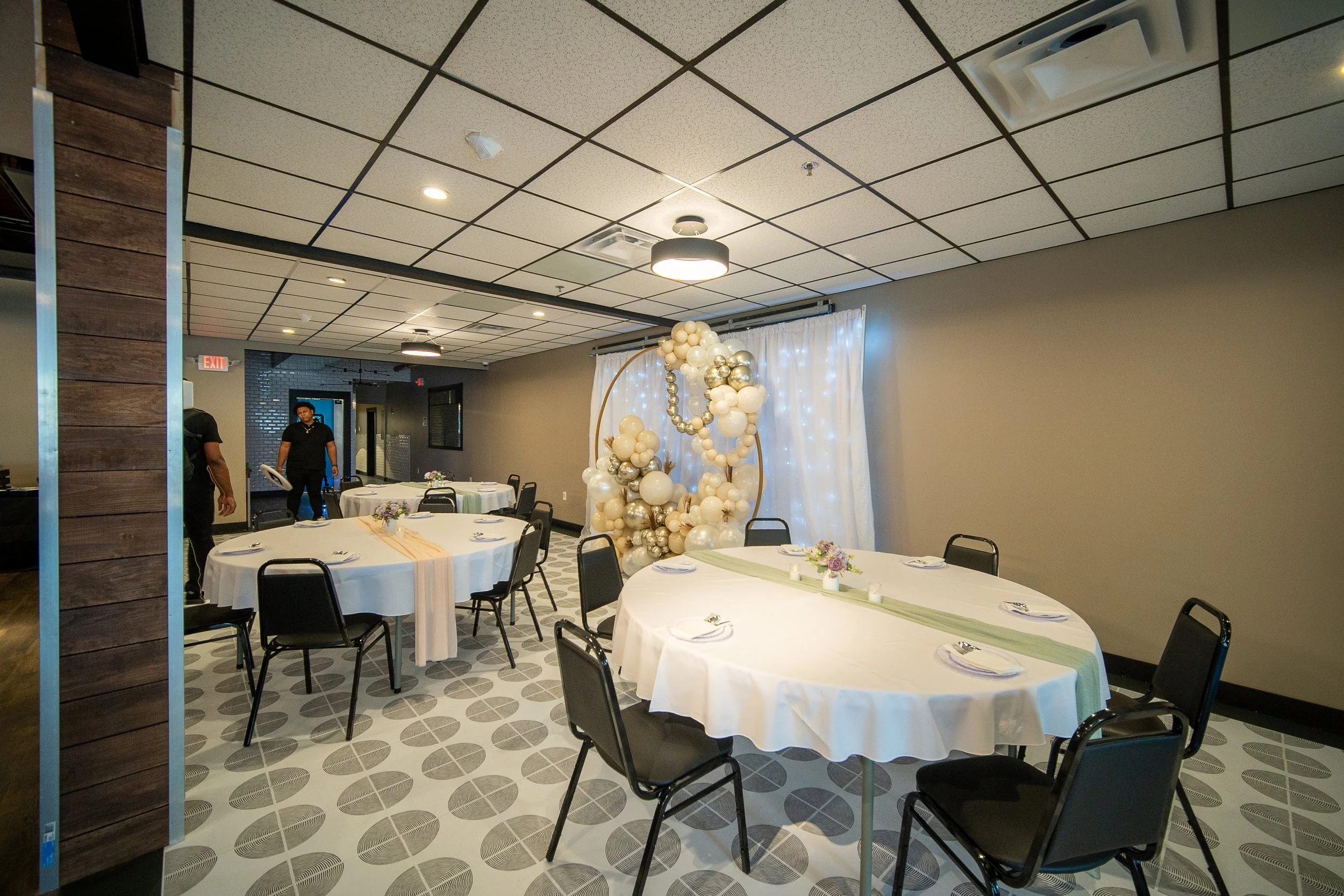 Decorated event room with round tables, white tablecloths, chairs, a balloon arch, and soft lighting.