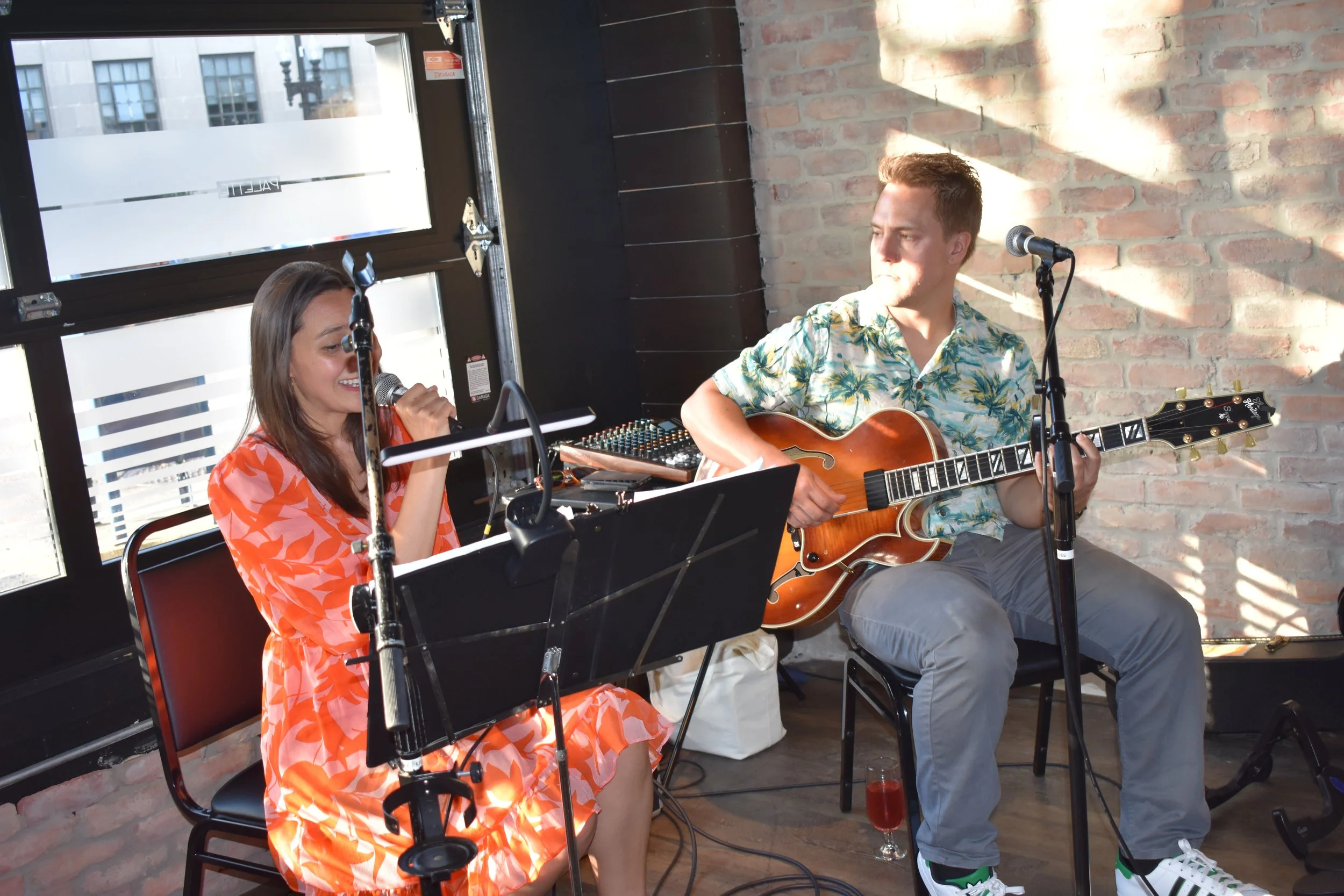 A woman in an orange dress sings into a microphone while a man in a floral shirt plays an electric guitar. They are seated inside a room with brick walls and large windows, performing with microphones and music stands.