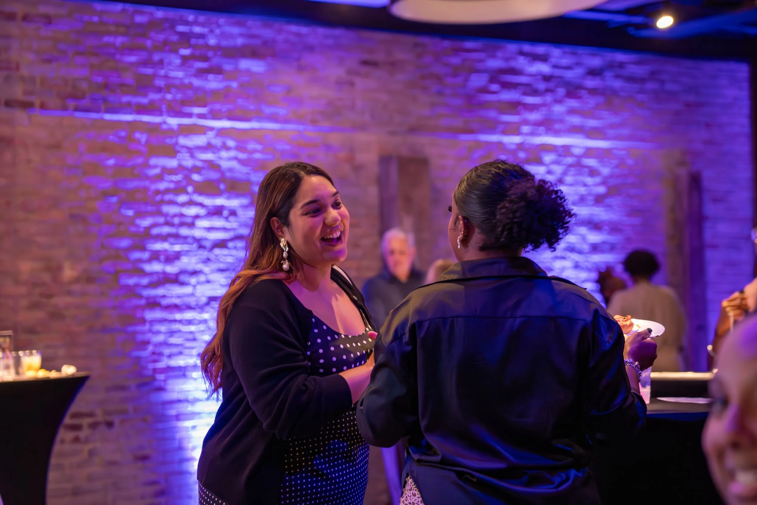 Two women are talking and laughing at a social event in a venue with brick walls and purple lighting. One woman has long brown hair, wearing earrings and a black outfit with white polka dots. The other woman has her hair styled in a curly updo, weari