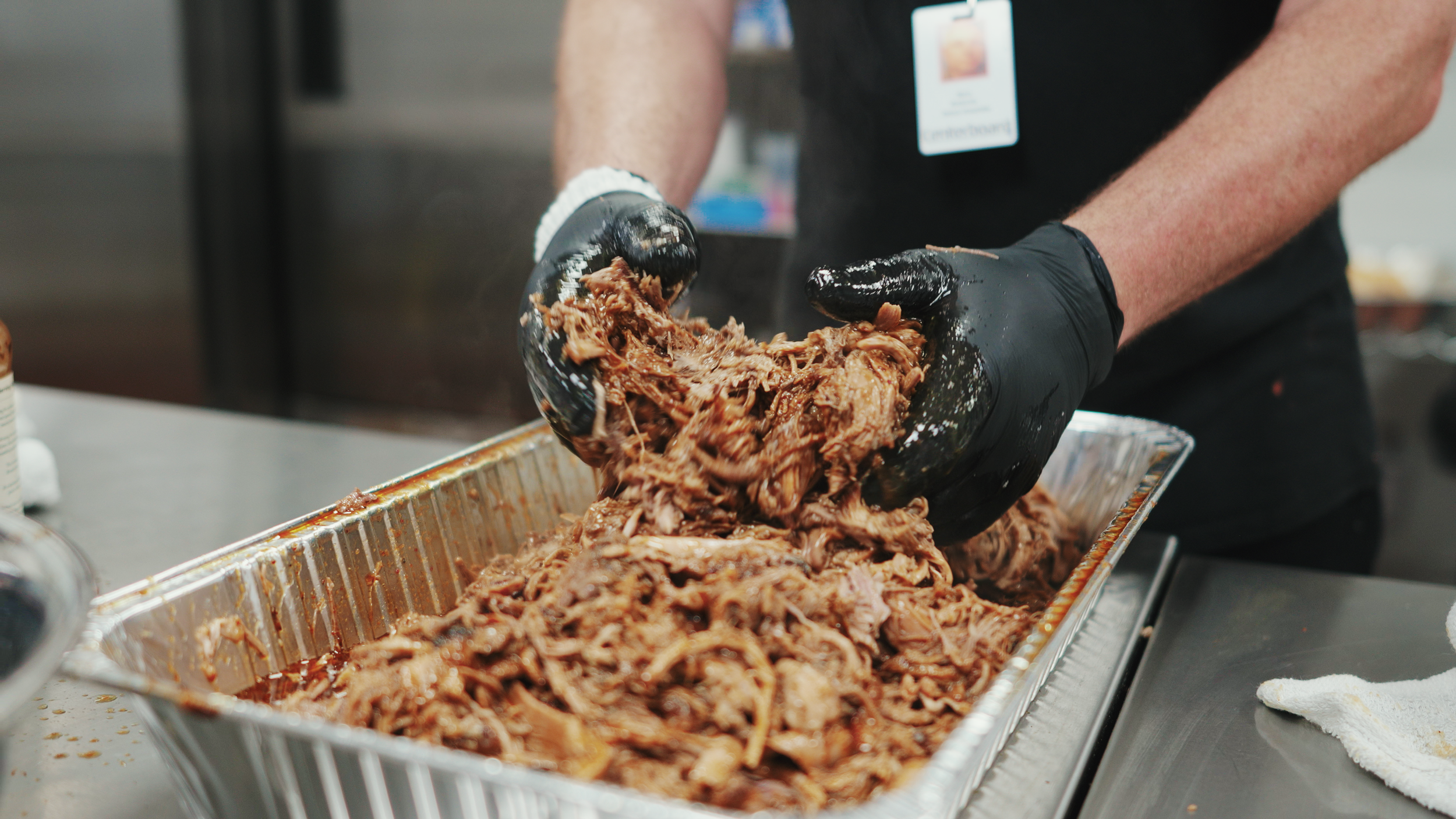 Person wearing gloves preparing pulled pork in a metal tray.