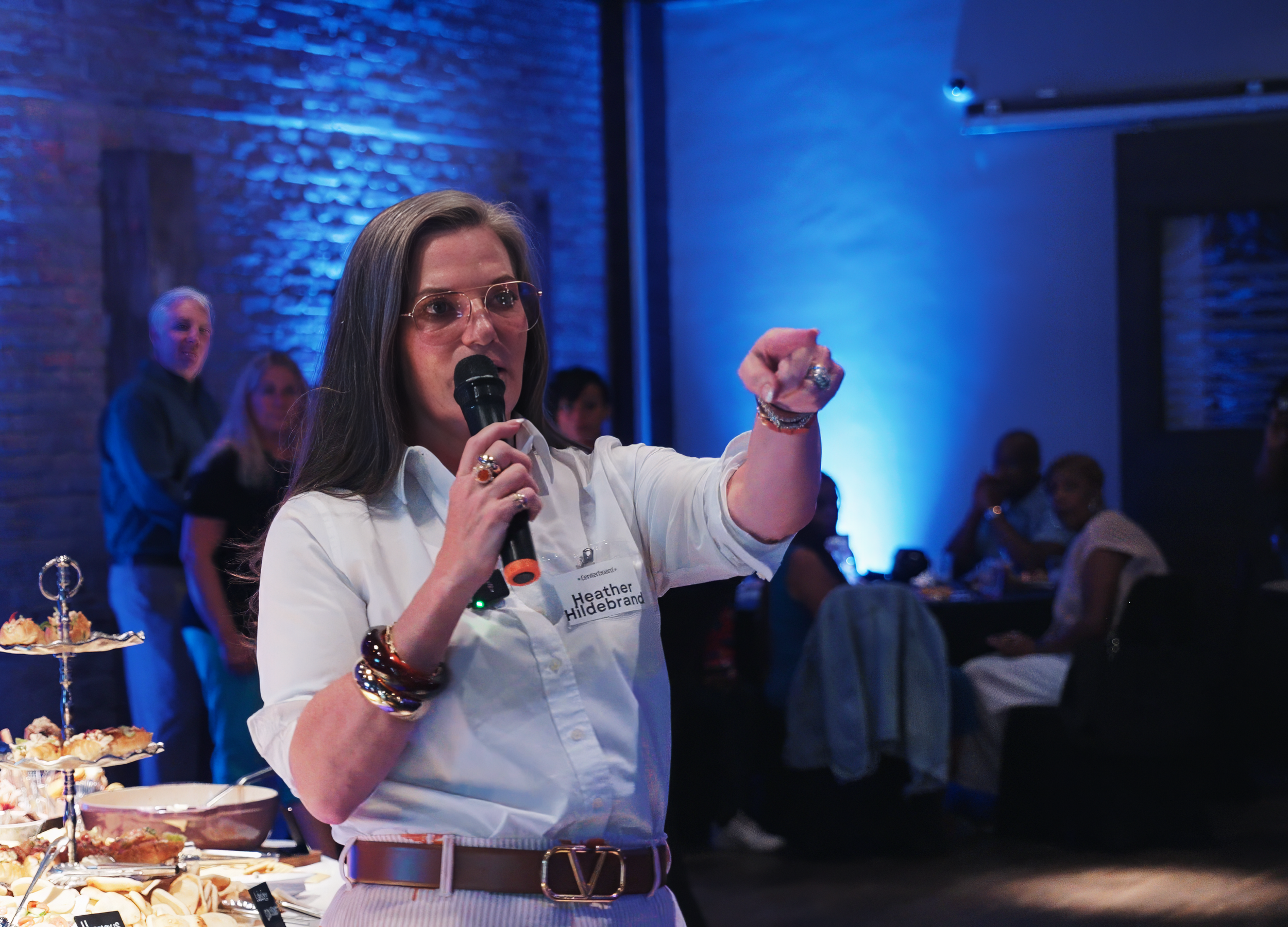A woman with long brown hair wearing glasses, a white shirt, and a brown belt, is speaking into a microphone at a social event. She is gesturing with her right hand while standing near a table with food. There are people sitting at tables and standin