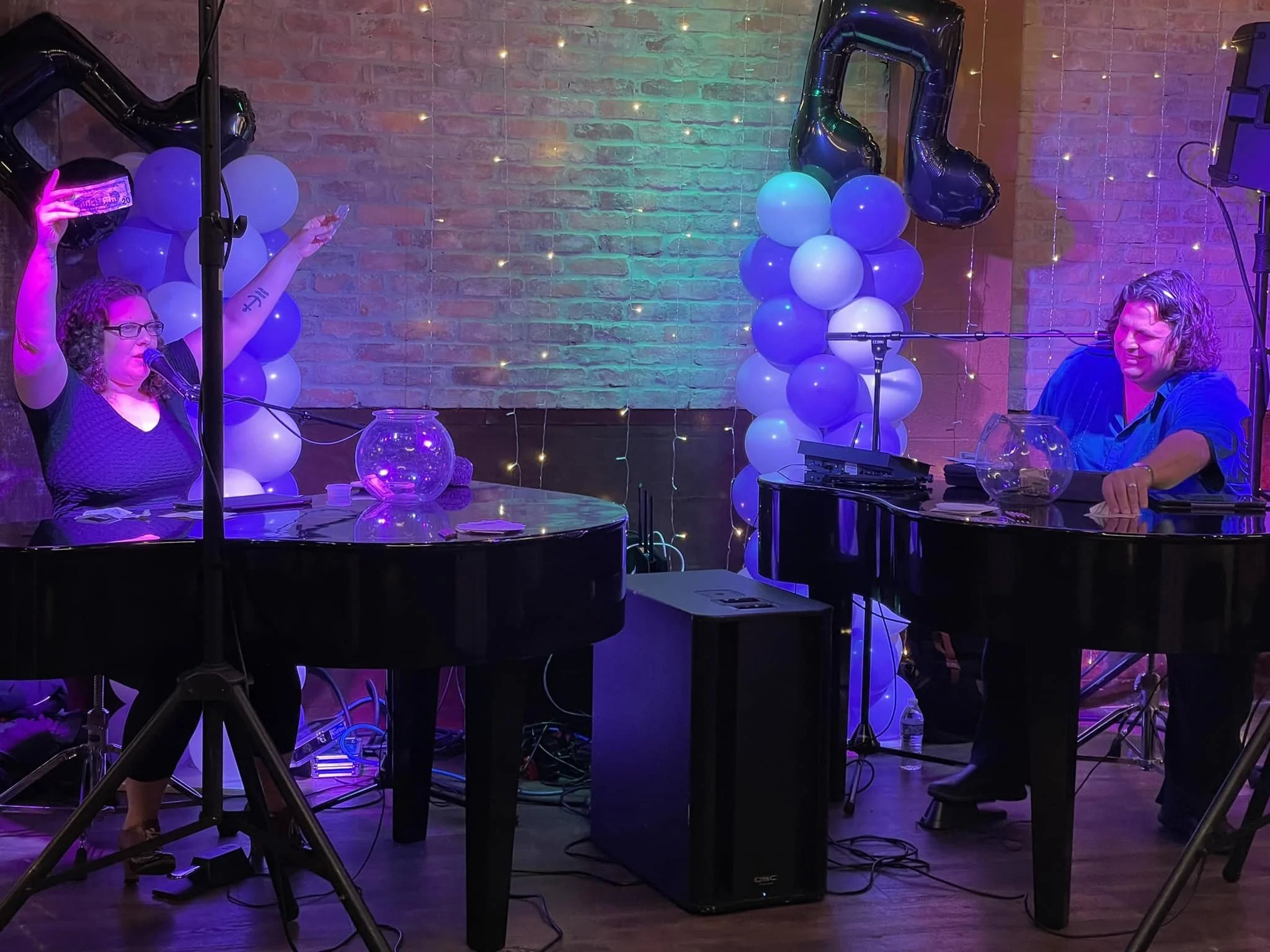 Two people seated at adjacent grand pianos, surrounded by balloons shaped like musical notes and clusters of purple and white balloons. One person raises their arm as they speak into a microphone. The setting includes brick walls and decorative strin
