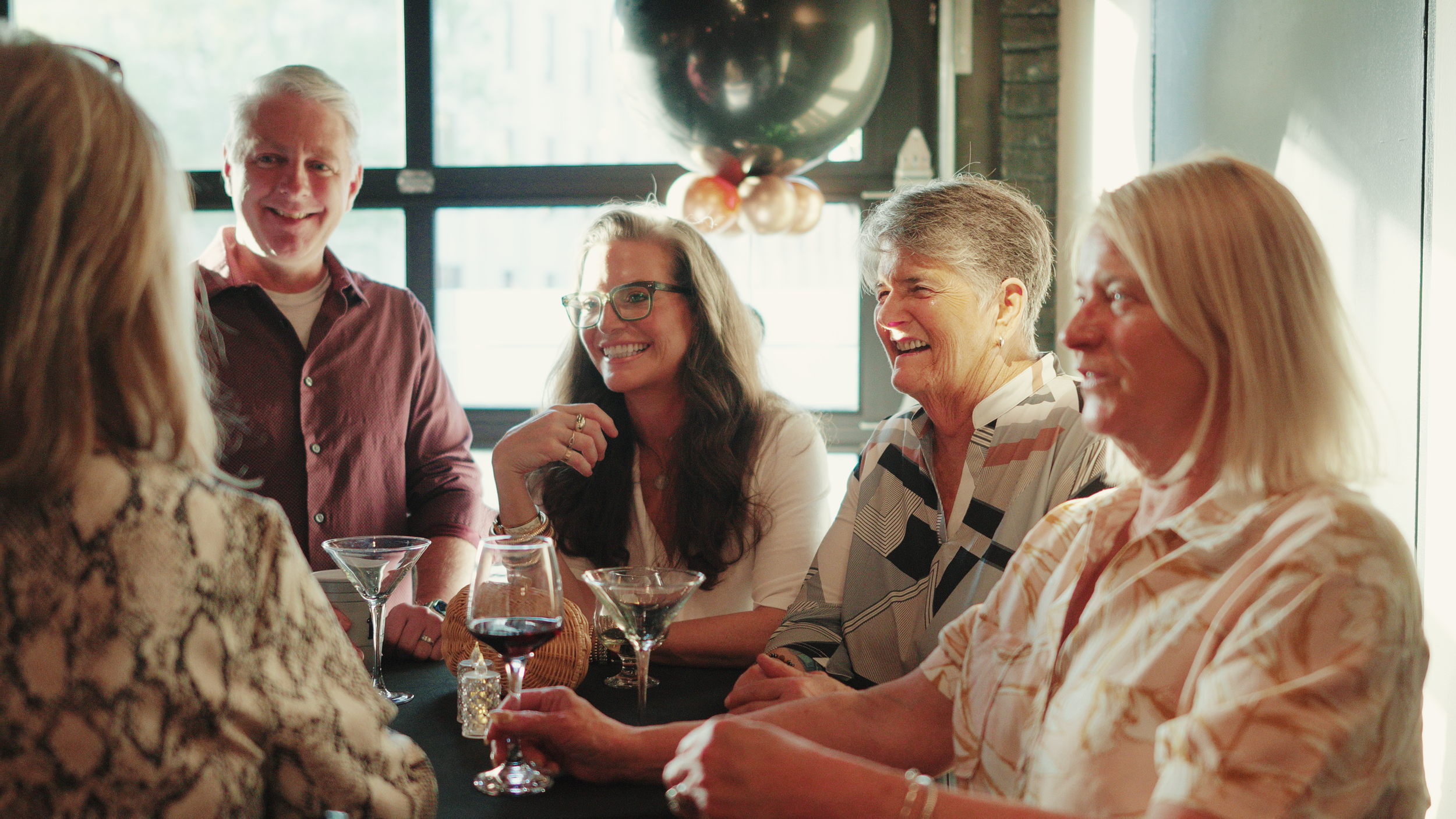 Group of people laughing and talking around a table with drinks at a social event.