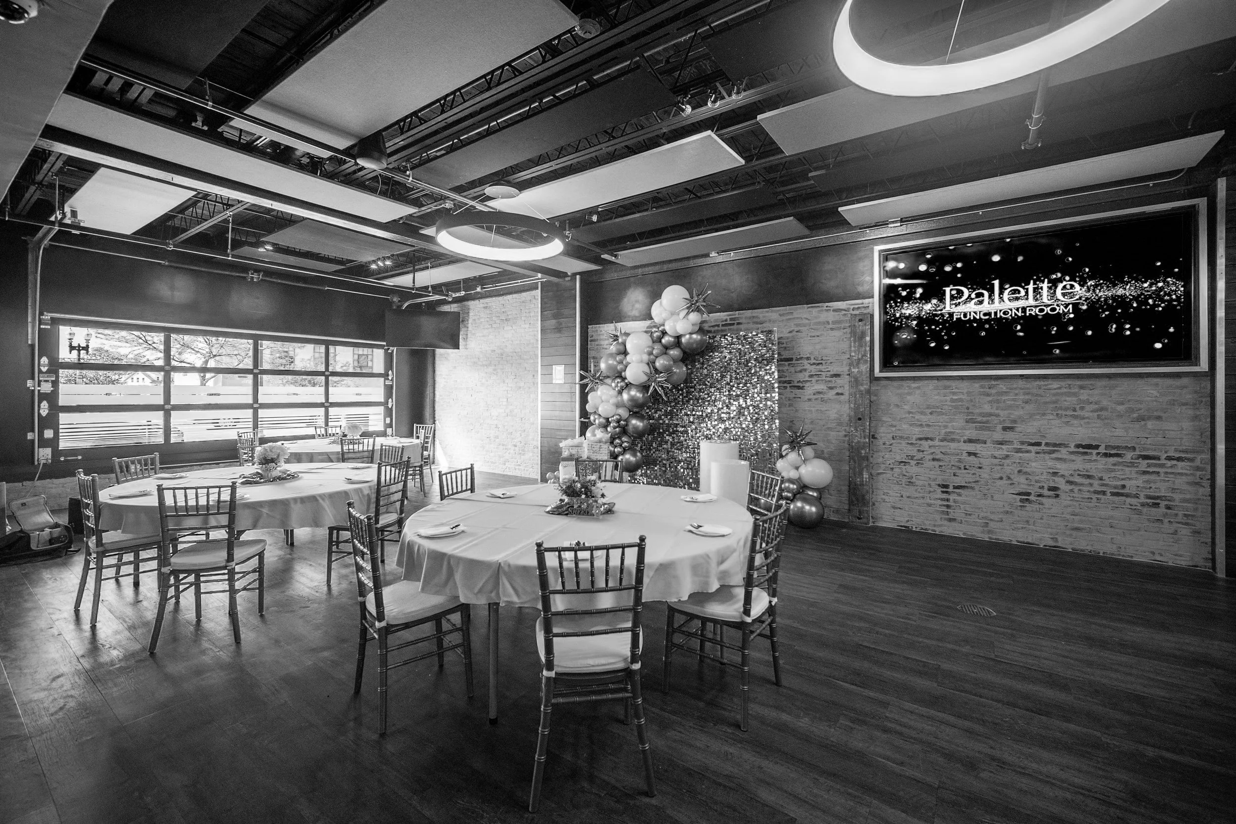 Black and white image of an empty event room with round tables, chairs, tablecloths, and a decorative balloon arrangement; a sign reads 'Palette Function Room.'
