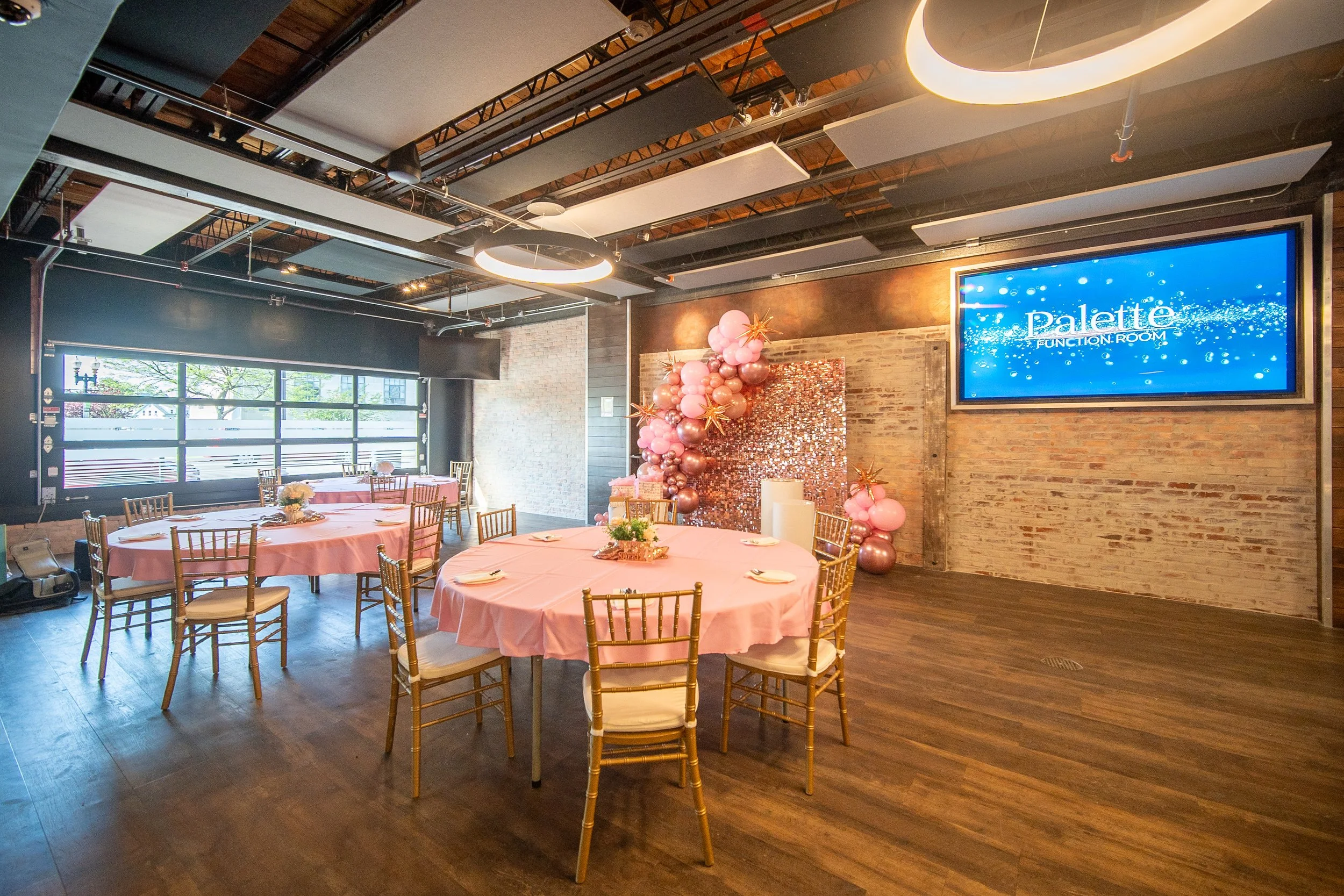 Event venue with round tables covered in pink tablecloths, gold chairs, balloon decorations, a display screen, and wooden floors.