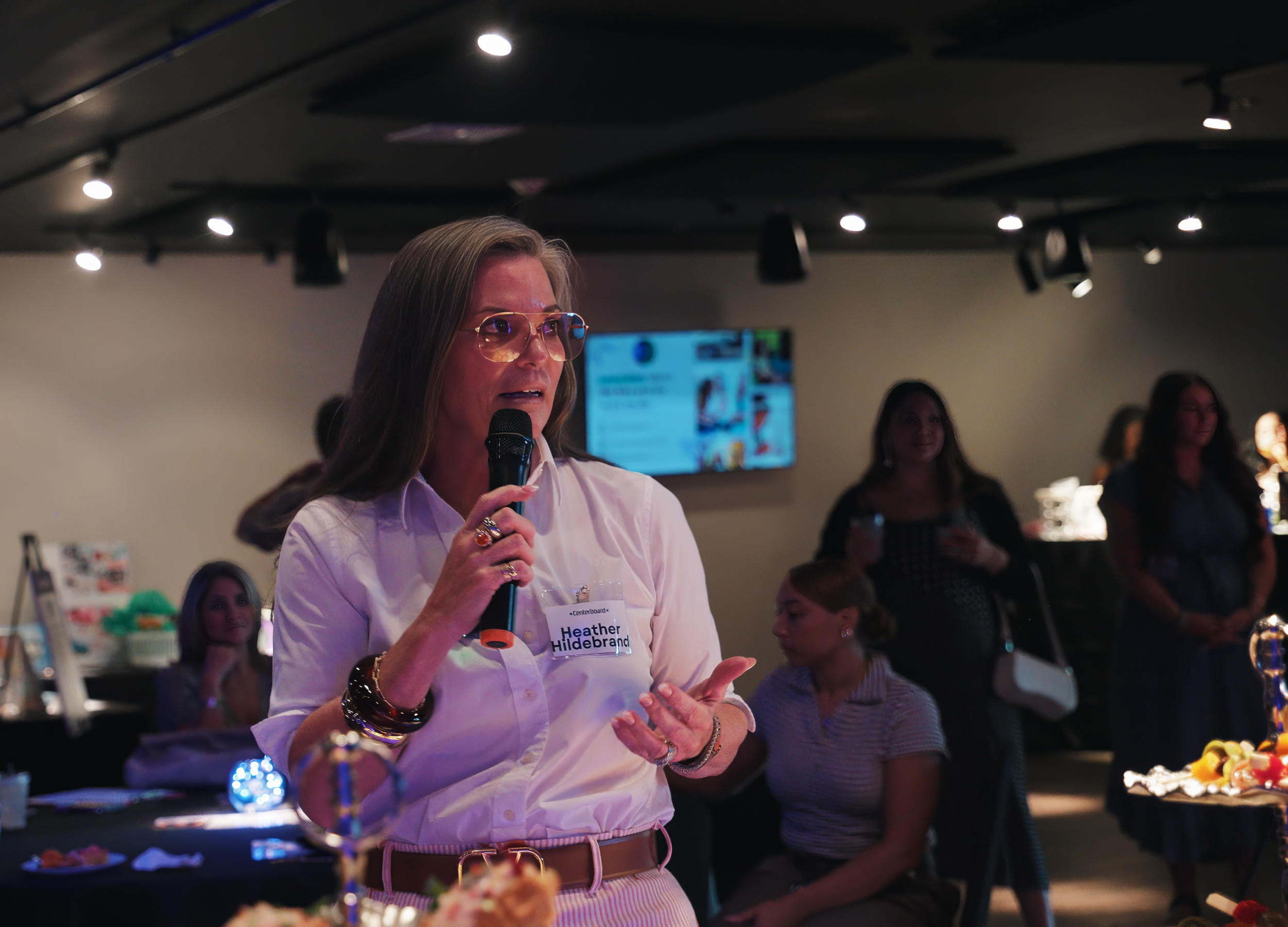 A woman wearing glasses, a white shirt, and a name tag labeled 'Heather Hildebrand' is holding a microphone and speaking at an indoor event. Several women are in the background, some seated and some standing, with a display screen and decorated table