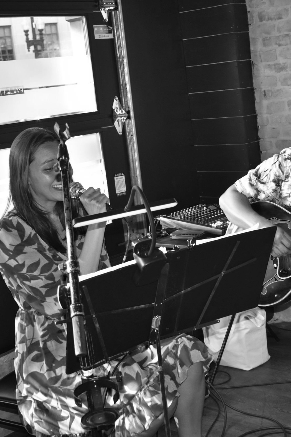 Black and white image of a woman singing into a microphone with a guitarist next to her, music stand and cables visible.