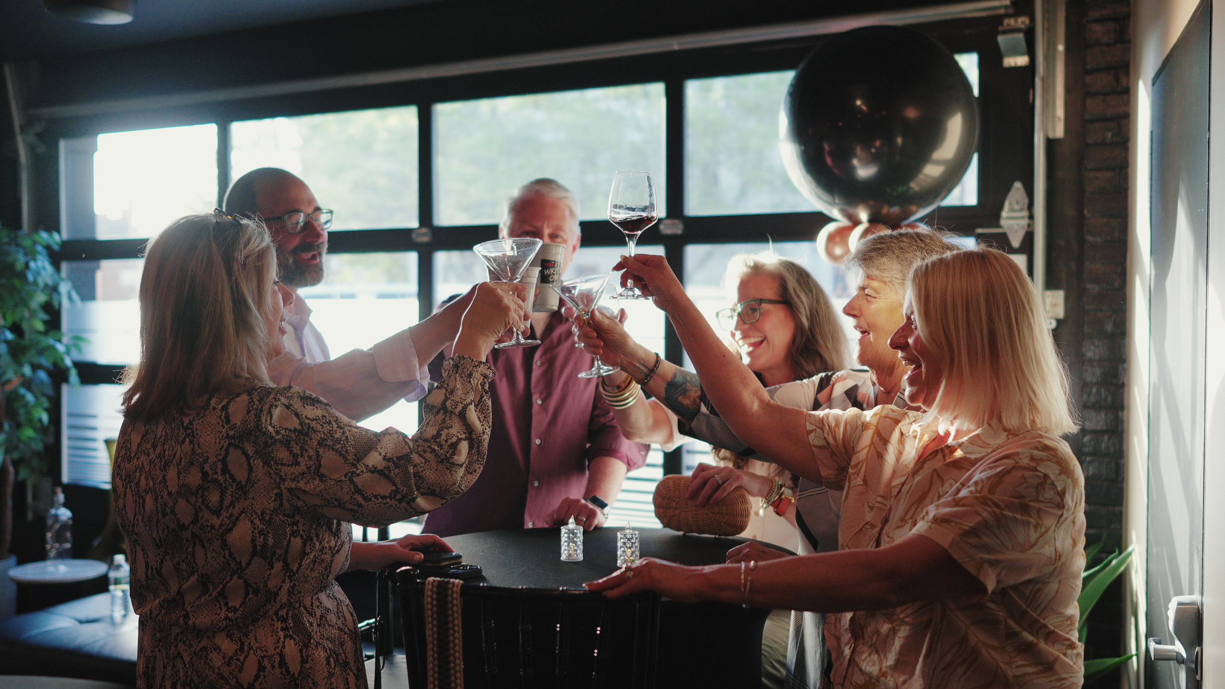 Group of six people raising glasses for a toast at a party, with a large balloon in the background.