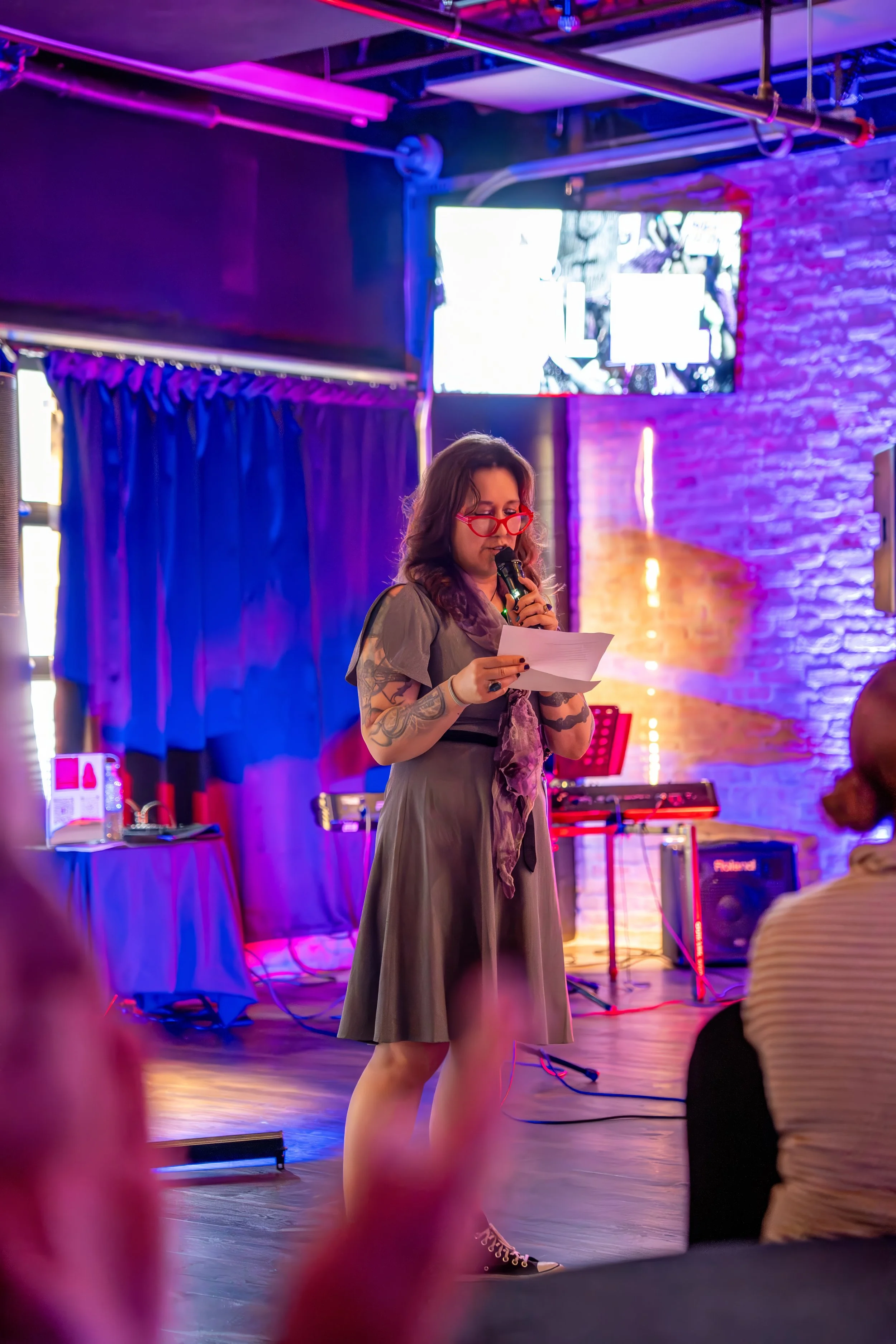 A woman with dark hair, red glasses, and tattoos on her arms reading from a paper while speaking into a microphone at a stage in a colorful indoor venue with purple and orange lighting, music equipment, and an audience.