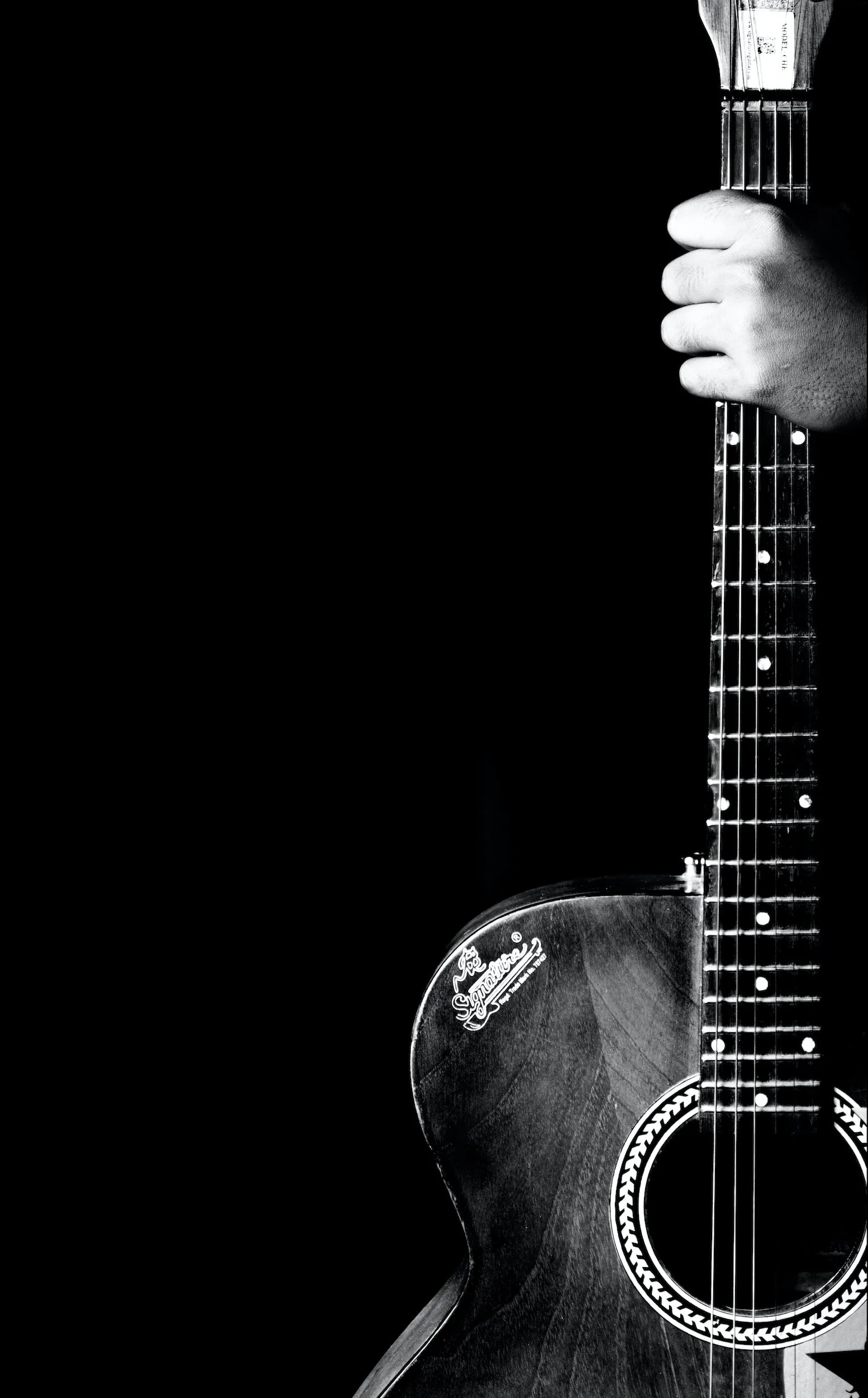 Close-up black and white photo of an acoustic guitar. The focus is on the body and neck of the guitar, with a hand resting on the side of the neck. The guitar has a sticker on the body that says 'Joe's' with some other text, and a patterned sound hole ring.