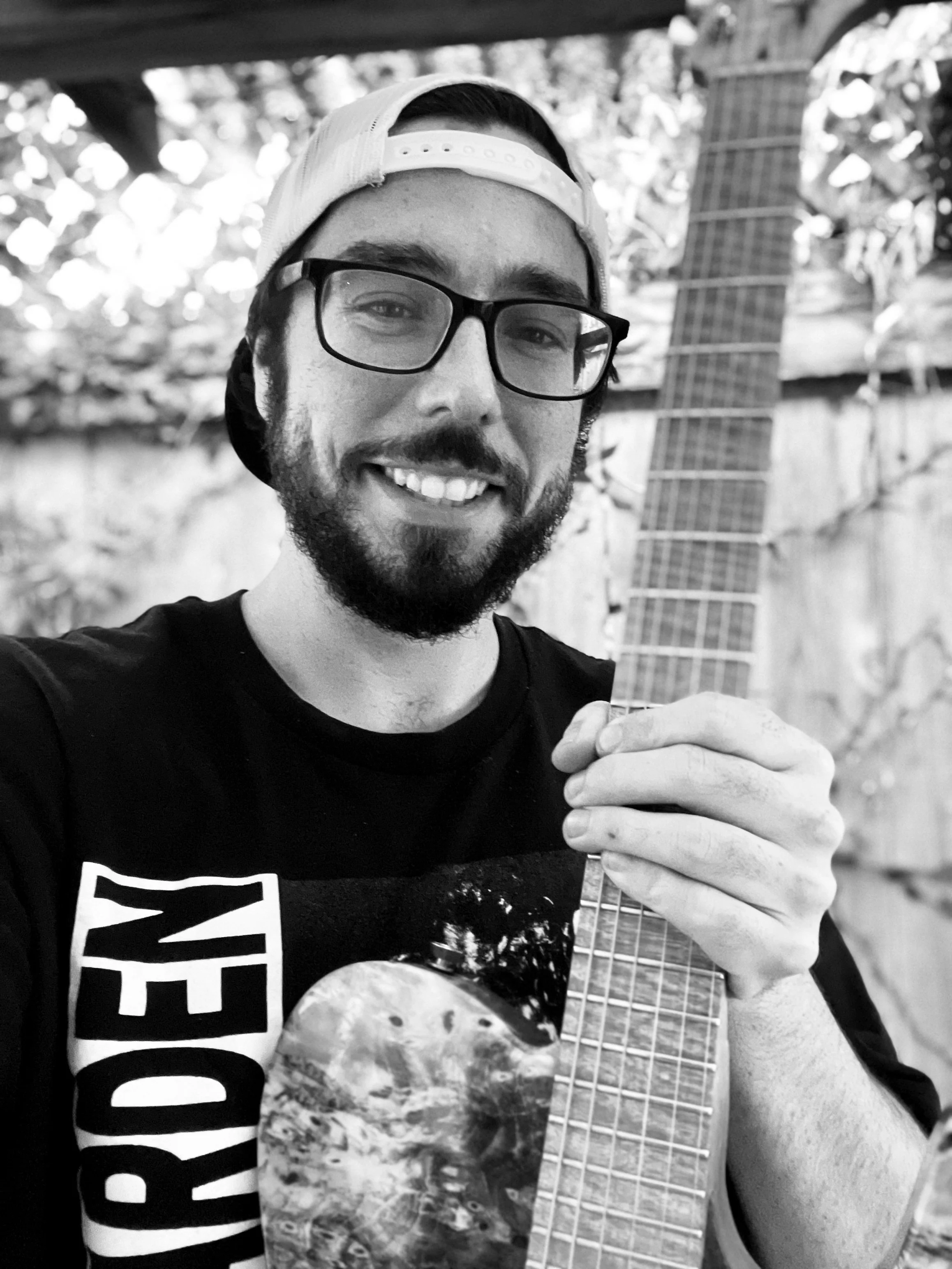 A young man with glasses and a baseball cap smiling while holding an electric guitar, standing outdoors in front of trees and a wooden structure.