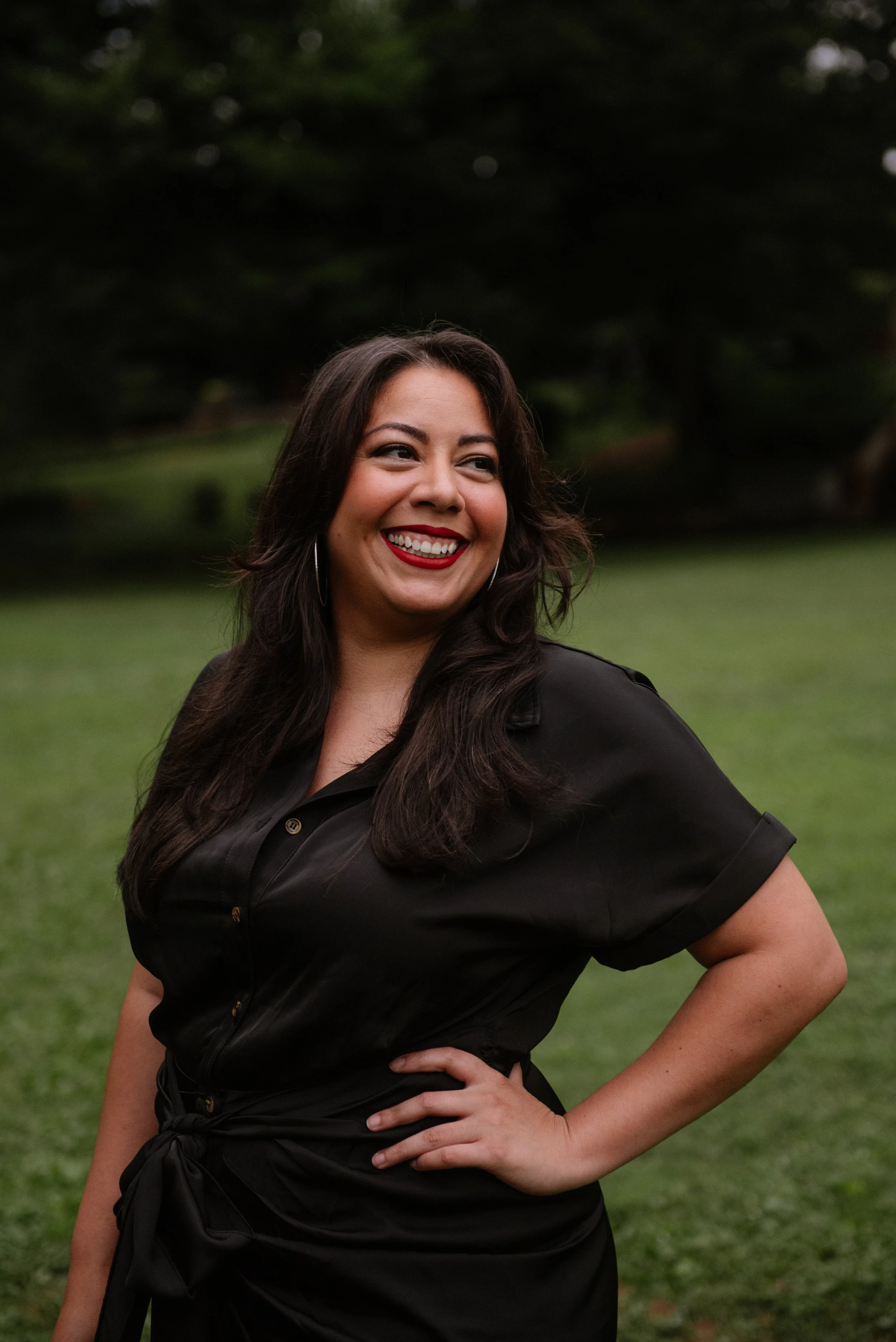 A woman with long dark hair, wearing a black dress with rolled-up sleeves, standing outdoors on grass with trees in the background. She is smiling, showing her teeth, and has her hand on her hip.