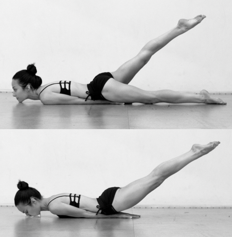 Black and white photo of a woman in a yoga pose on the floor, with one leg lifted and extended straight behind her, in a plank position.