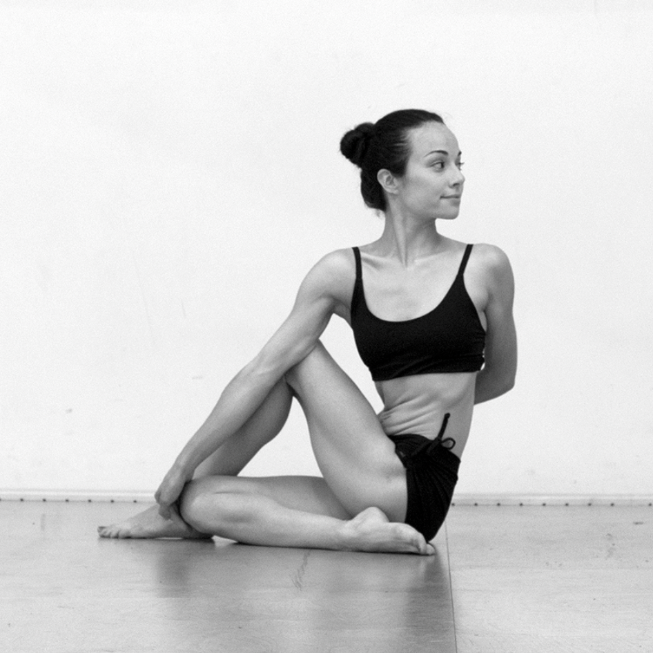 A woman sitting on the floor in a yoga pose against a plain wall.