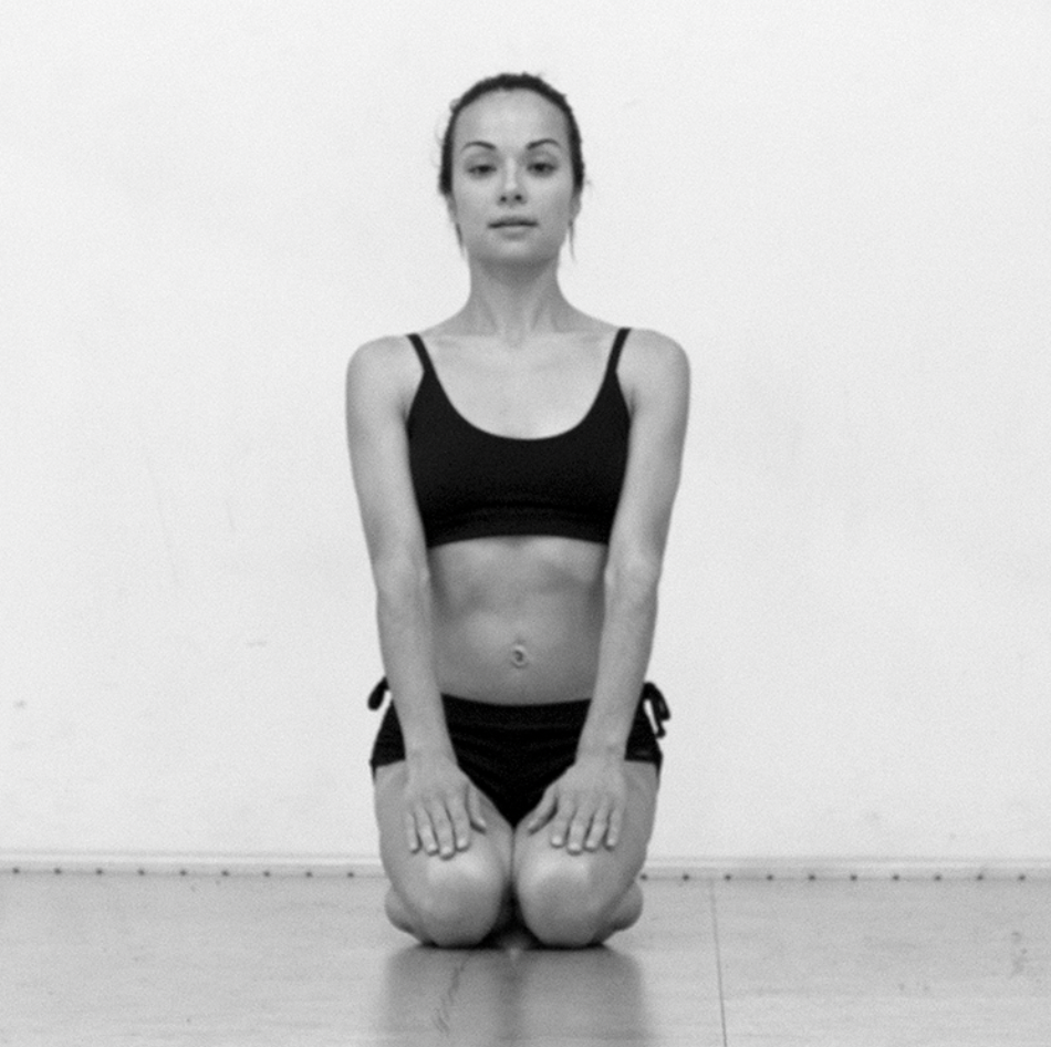 A woman in a black sports bra and shorts kneeling on the floor with her hands on her thighs, facing forward in front of a plain wall.