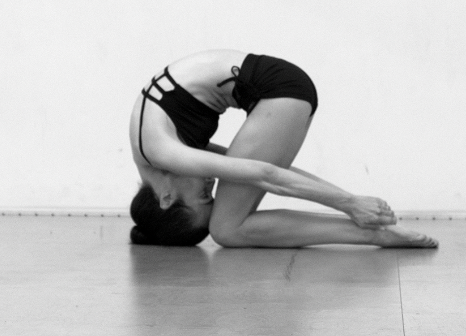 A woman in black shorts and a tank top practicing yoga in a head-to-kike pose, bending forward with arms extended and hands clasped behind her feet on a wooden floor.