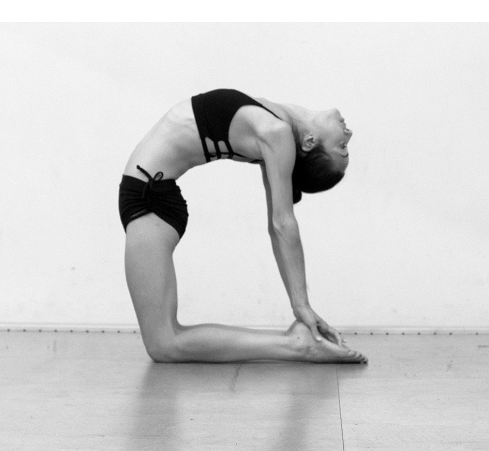 A woman practicing yoga in a black sports bra and shorts, performing a backbend pose on a wooden floor with a plain white wall background.