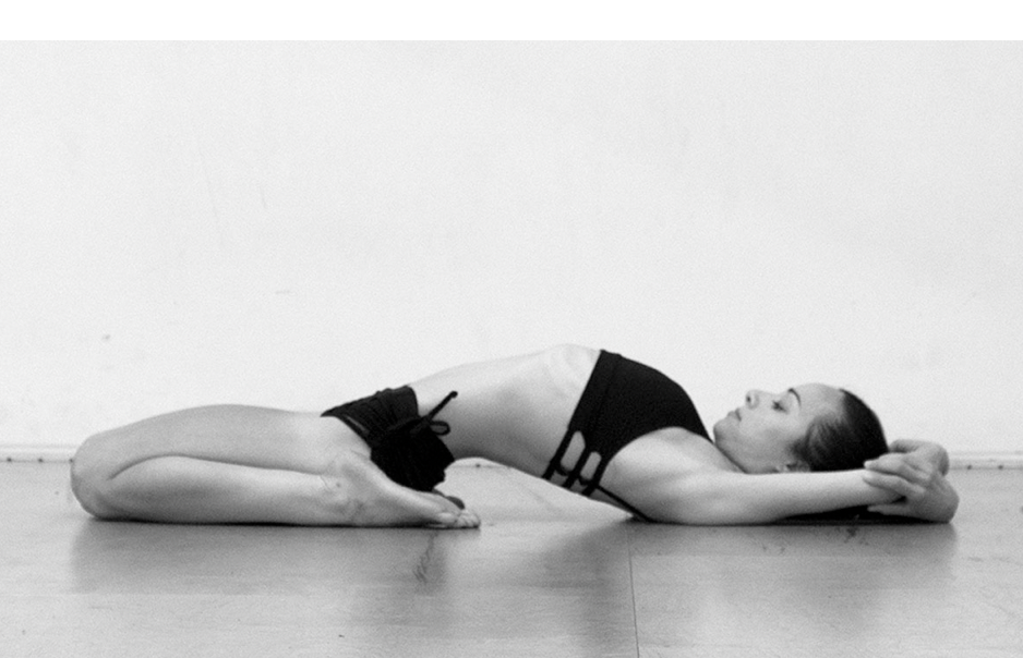 Woman in black workout attire lies on floor in yoga pose with arms extended above head and knees bent.