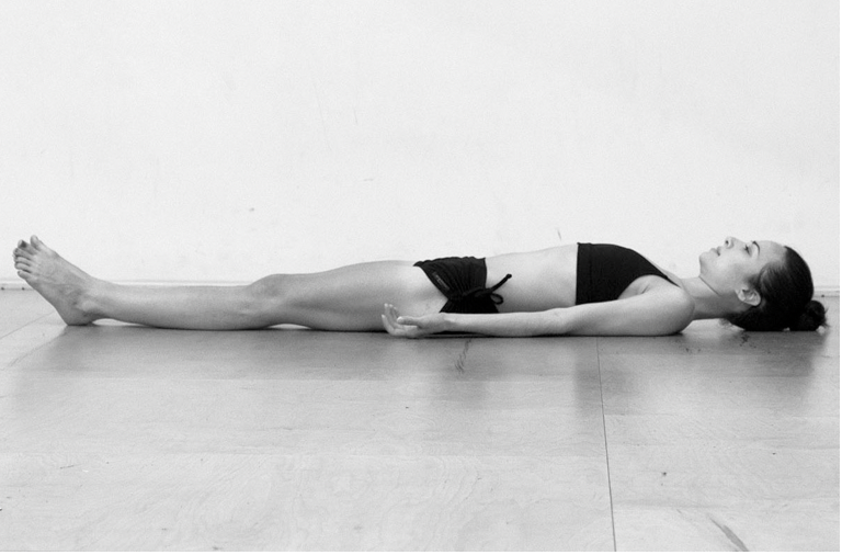 A woman lying on her back on a wooden floor, in a yoga or relaxation pose, wearing a sleeveless top and shorts.