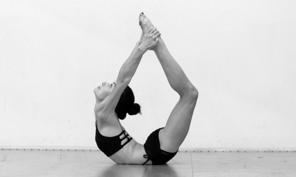 A woman in black workout clothing performing a yoga pose on a smooth floor, against a plain white wall.
