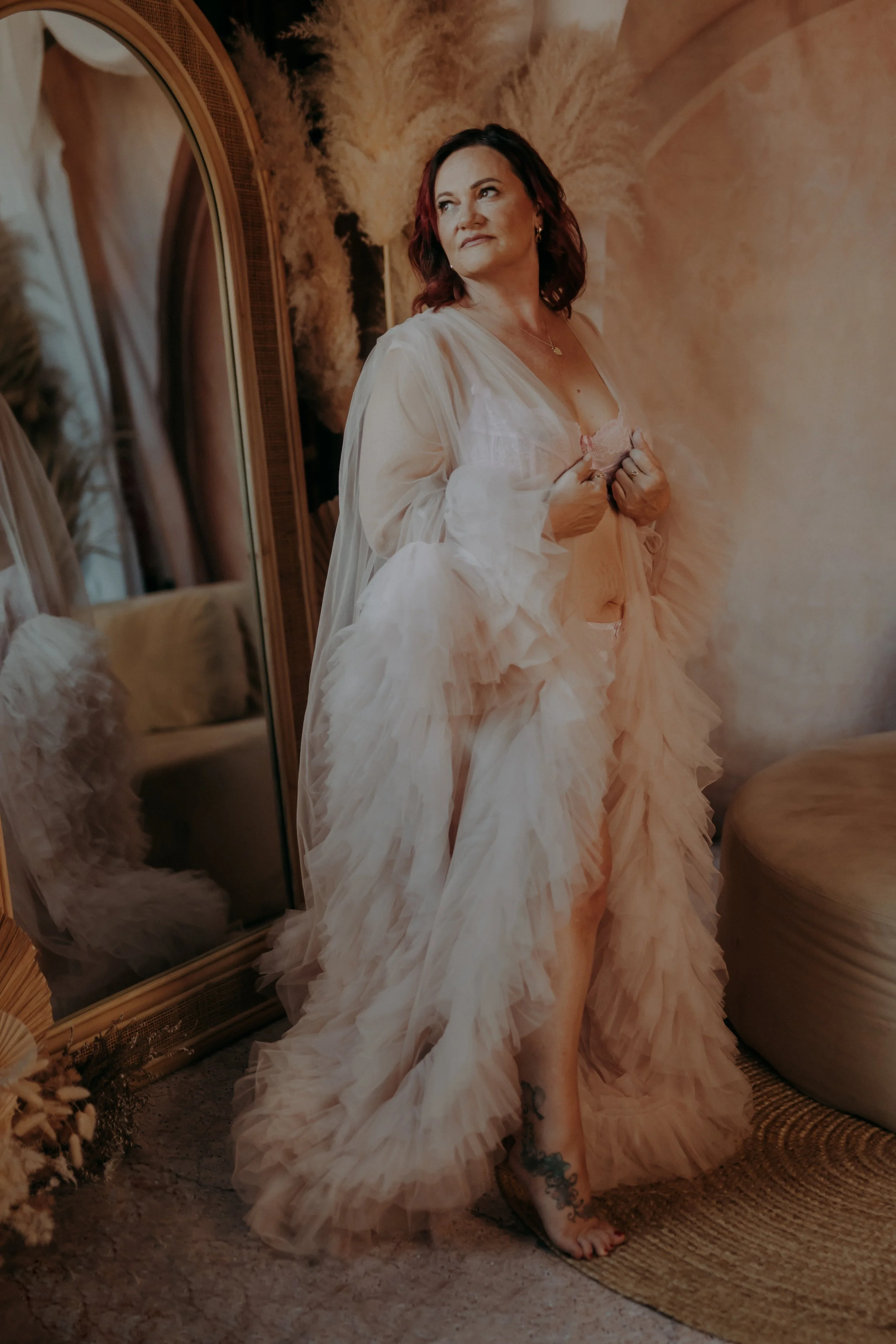 A woman with dark red hair and a tattoo on her foot stands in front of a mirror, wearing a flowing, ruffled, cream-colored gown, adjusting her top with a thoughtful expression.