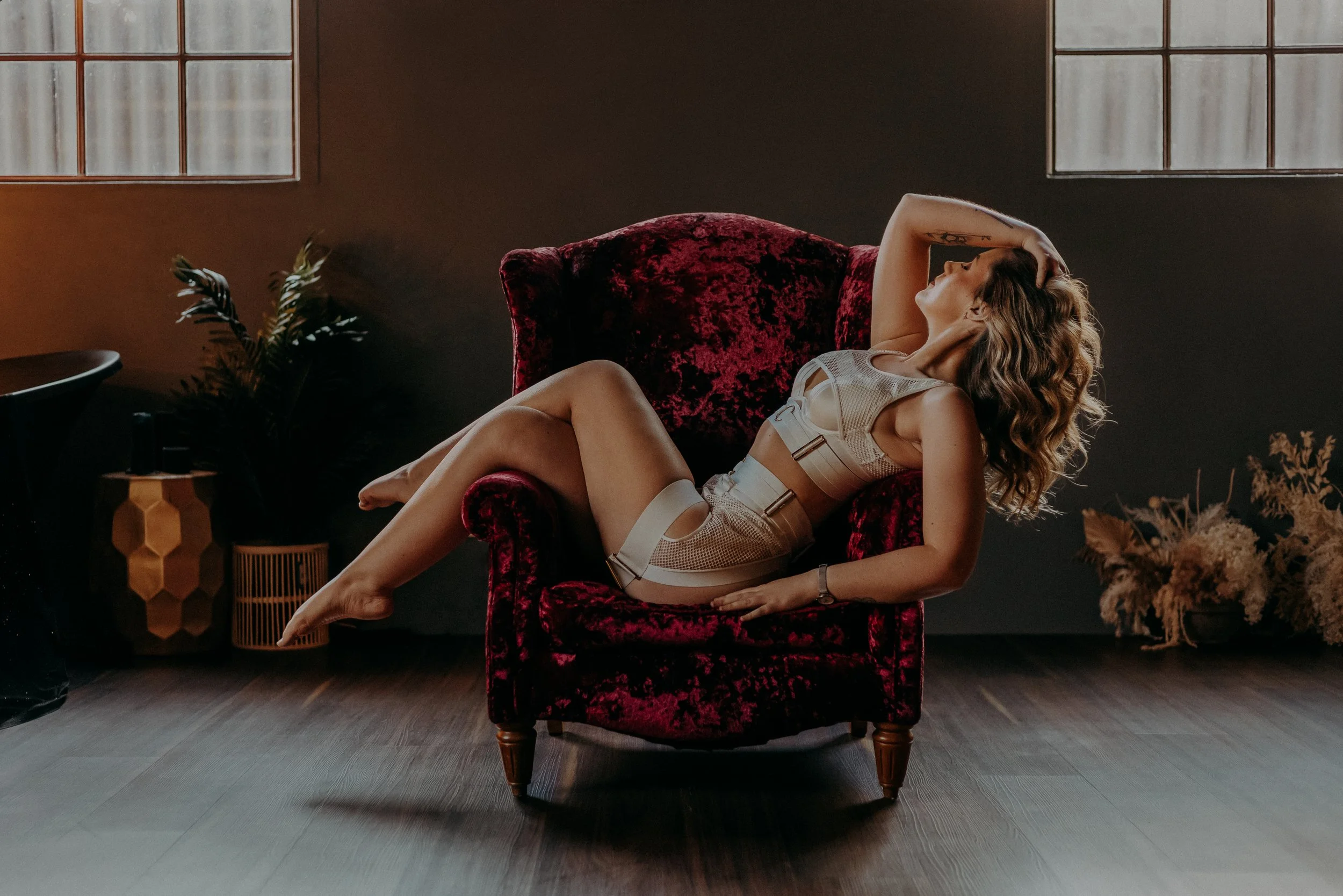 A woman with wavy hair lying on a plush red velvet armchair in a dimly lit room, wearing a beige outfit with cutouts and straps.