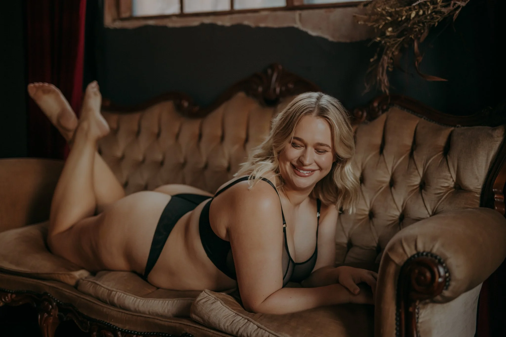 A woman in black lingerie lying on her stomach on a vintage gold sofa, smiling with her eyes closed, in a dimly lit room with dark walls and red curtains.