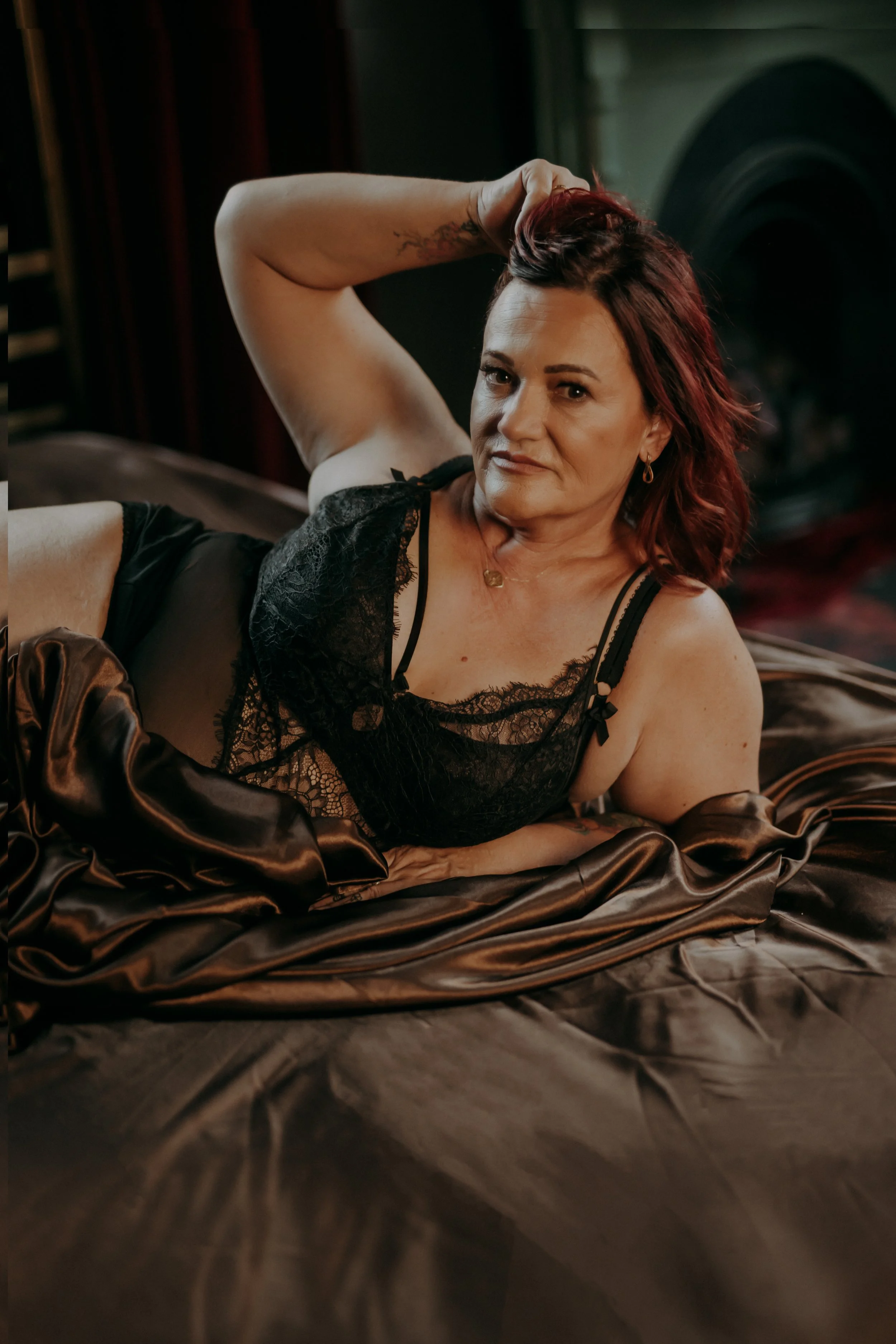 A woman with red hair lying on a satin bed in black lace lingerie, posing with one hand behind her head and looking at the camera.