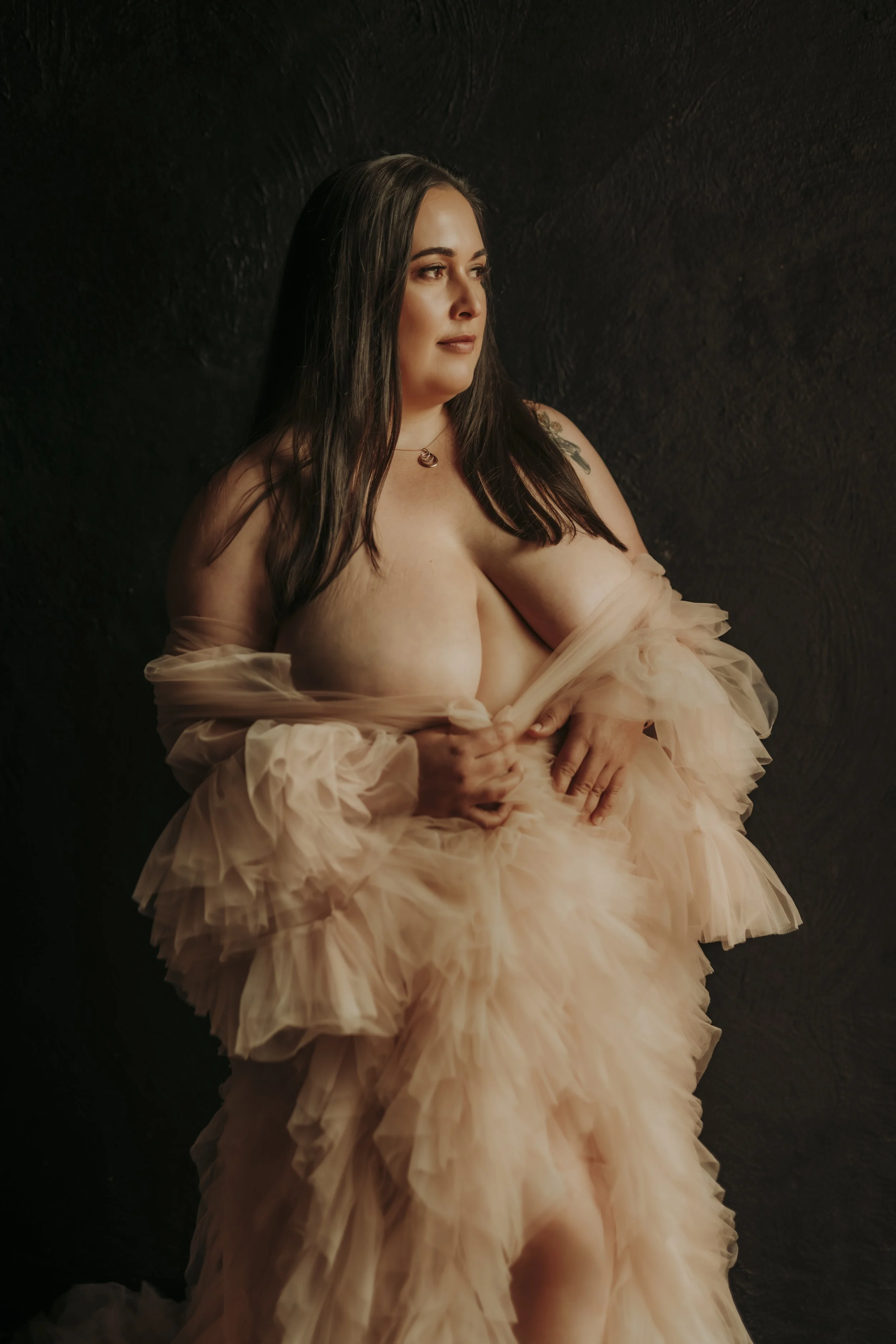 A woman with dark hair, tattoos, and a red velvet dress sitting on the floor, wearing a floral crown with red roses, in an artistic indoor setting.