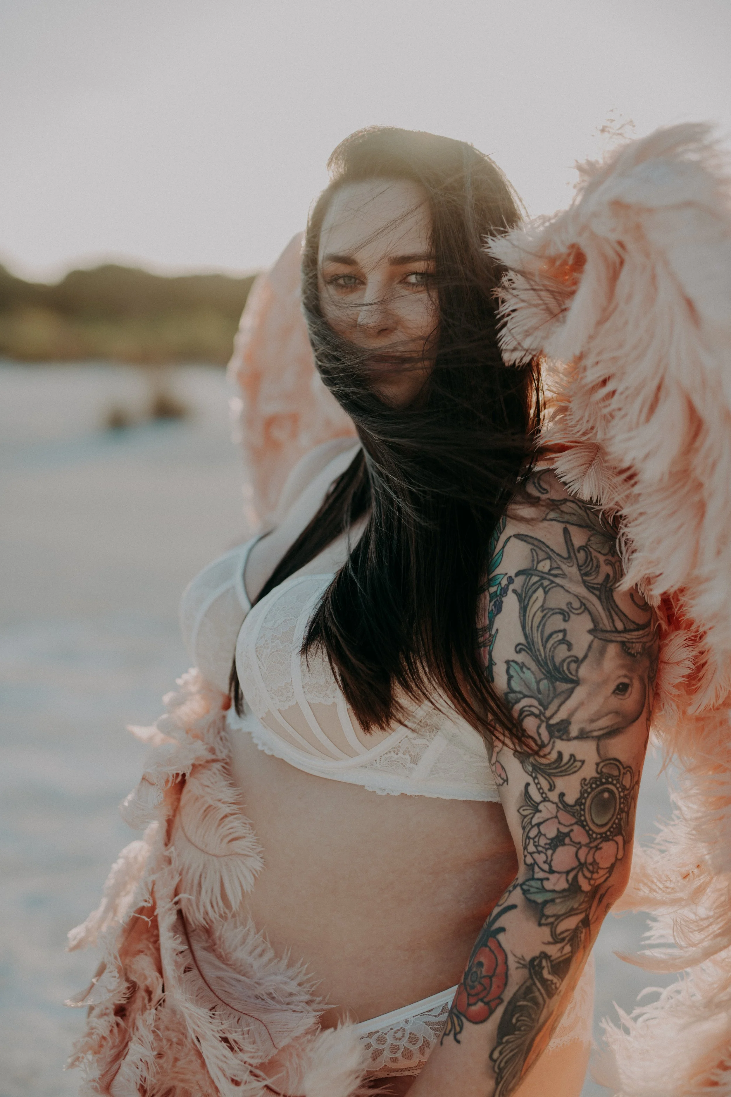 A woman with long dark hair, tattoos on her arm, and wearing white lace lingerie, stands on a beach with a feathered pink garment, looking at the camera with the wind blowing her hair.